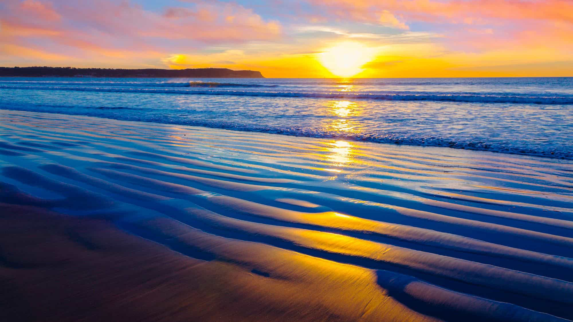 A golden sunset reflected in the wet rippled sand, creating a stunning interplay of light and shadow, with gentle waves rolling onto the shore.
