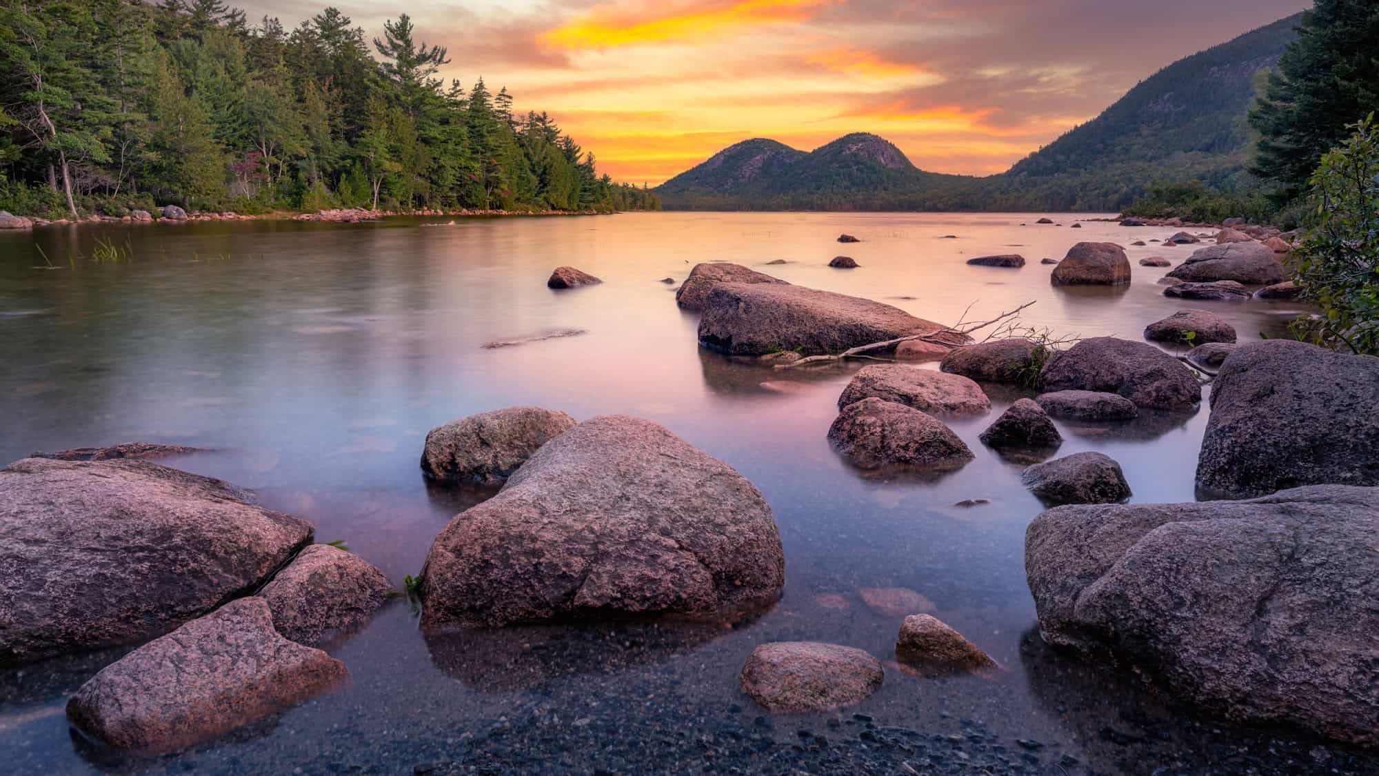 A calm pond dotted with large boulders at the shore, surrounded by lush evergreen trees and two rounded hills in the distance under a colorful sunset sky.