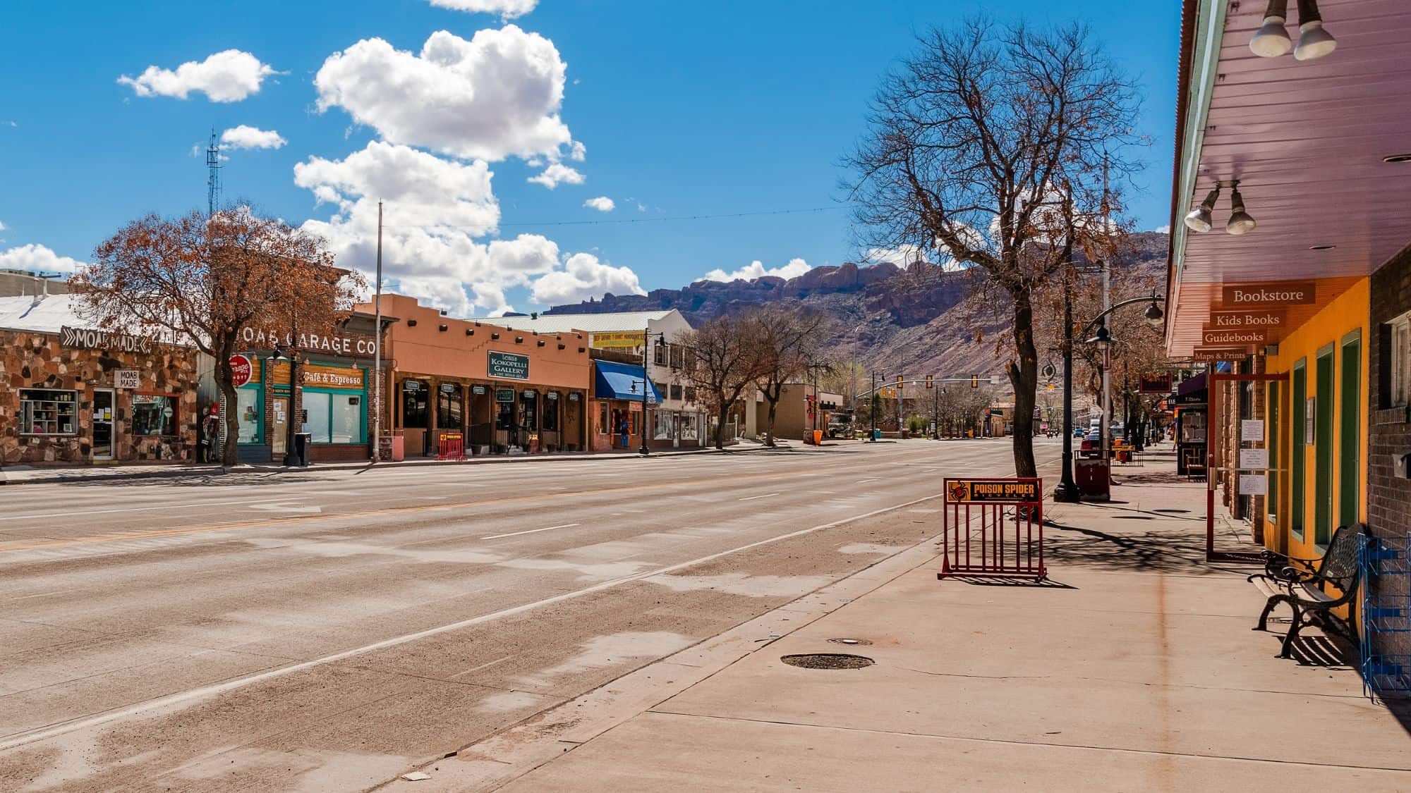 A quiet main street in Moab, Utah, with a mix of colorful storefronts and barren trees, set against a dramatic backdrop of rugged desert cliffs under a bright blue sky.
