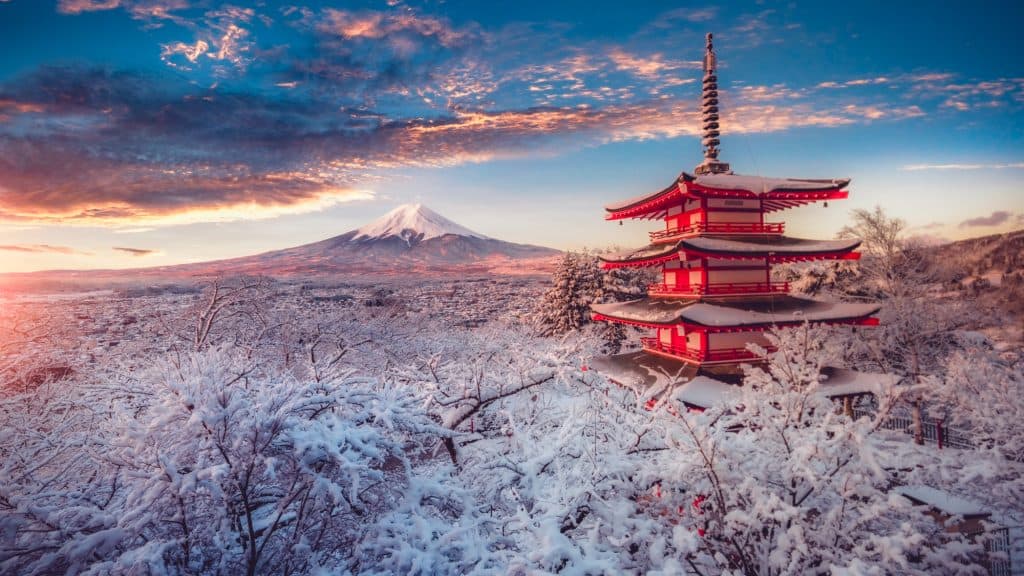 A breathtaking view of Mount Fuji framed by a red pagoda and snow-covered cherry blossom trees. The pastel-colored sky adds warmth to the cold winter scene.