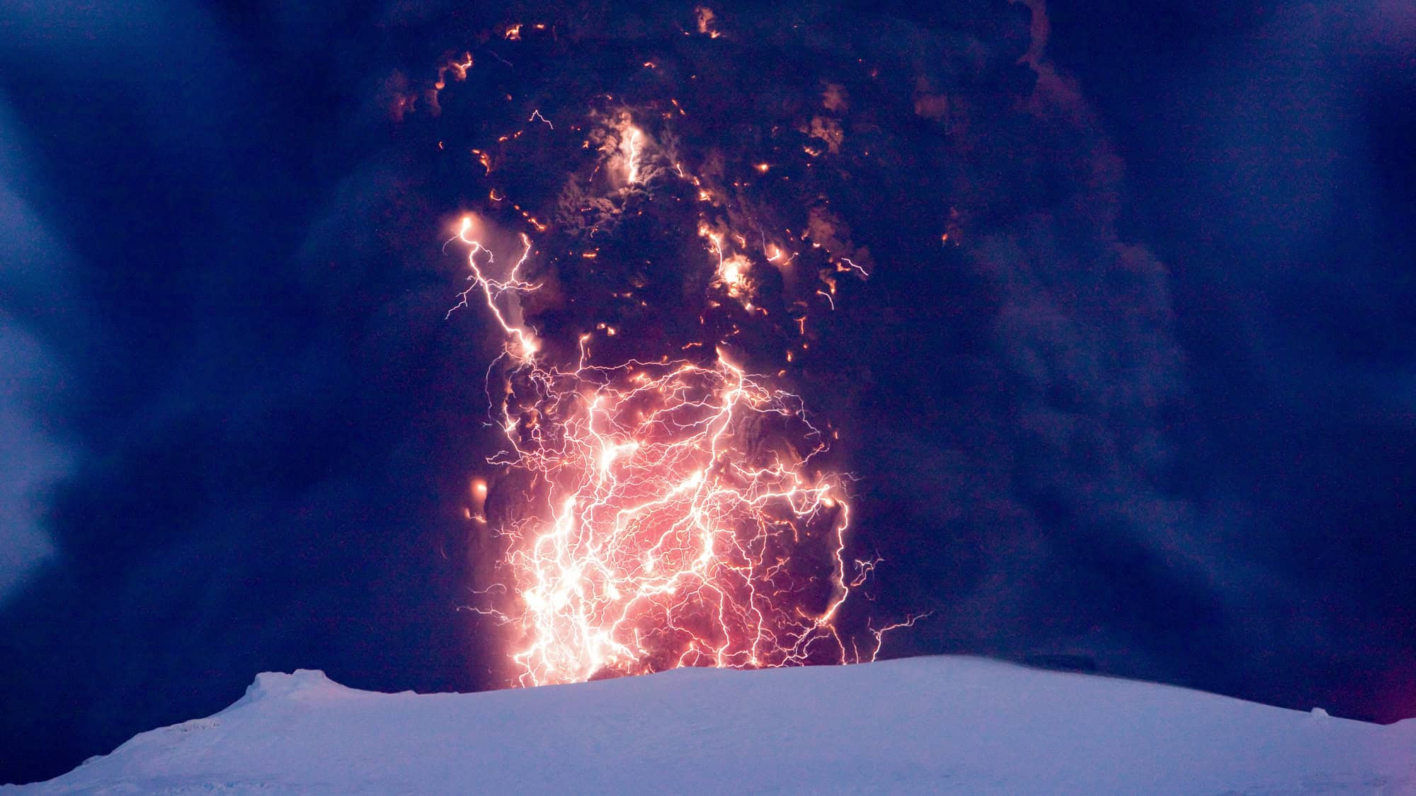 A massive volcanic eruption spews dark ash as bolts of lightning crackle through the plume against the night sky.