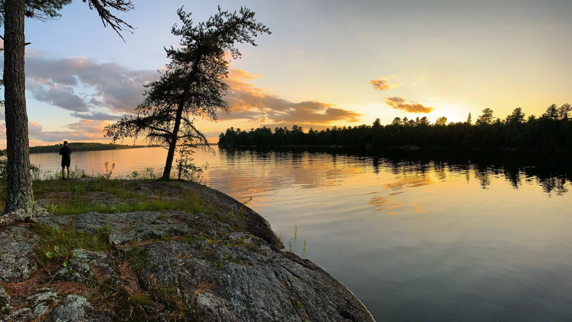 A peaceful sunset view at Voyageurs National Park with golden light reflecting off a calm lake, silhouetted trees along the shoreline, and a lone person standing near the rocky, mossy edge.