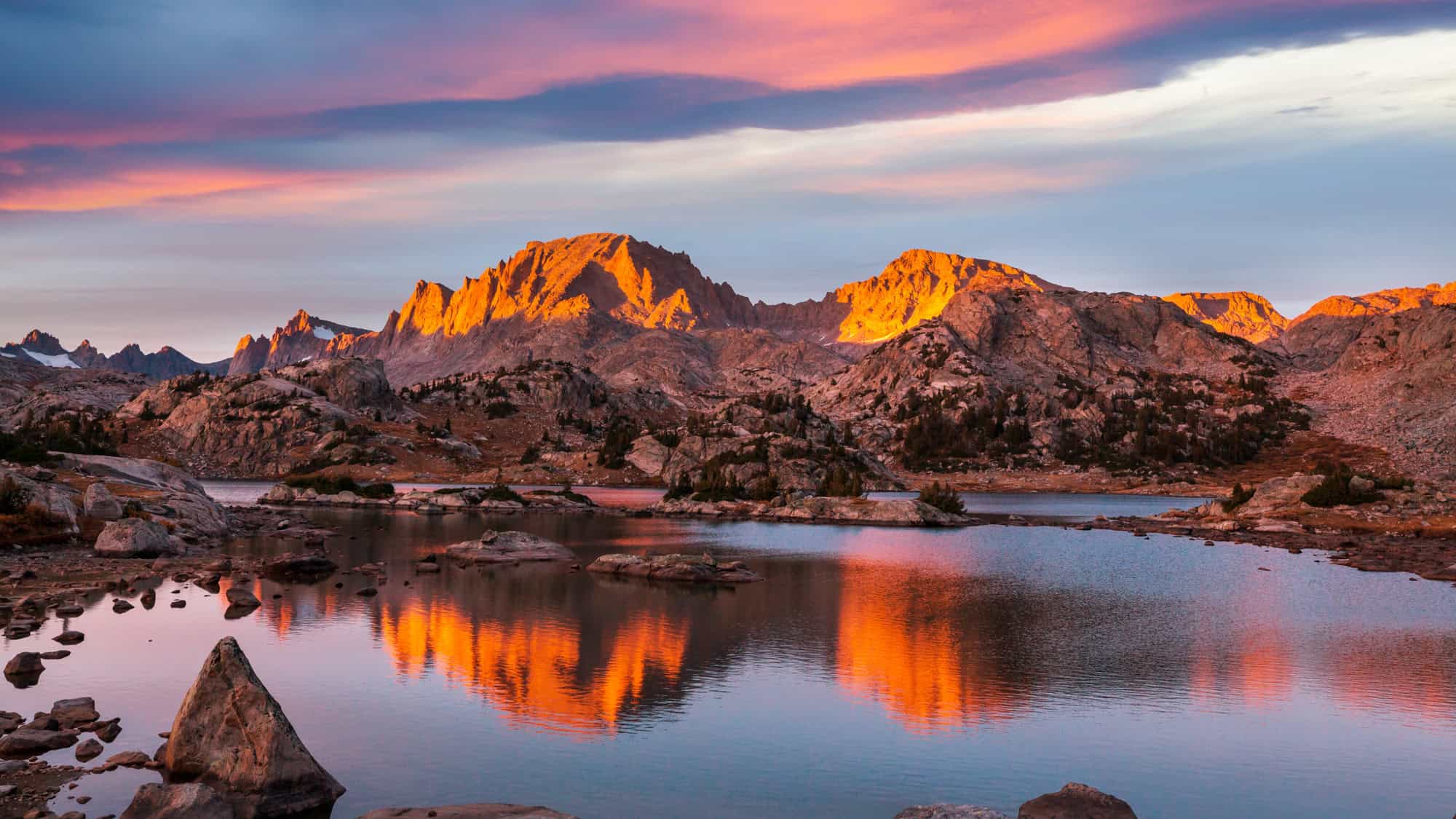 A glowing sunrise bathes the jagged granite peaks of Wyoming’s Wind River Range in warm orange light, with their reflection vividly mirrored in the calm, rocky alpine lake below.