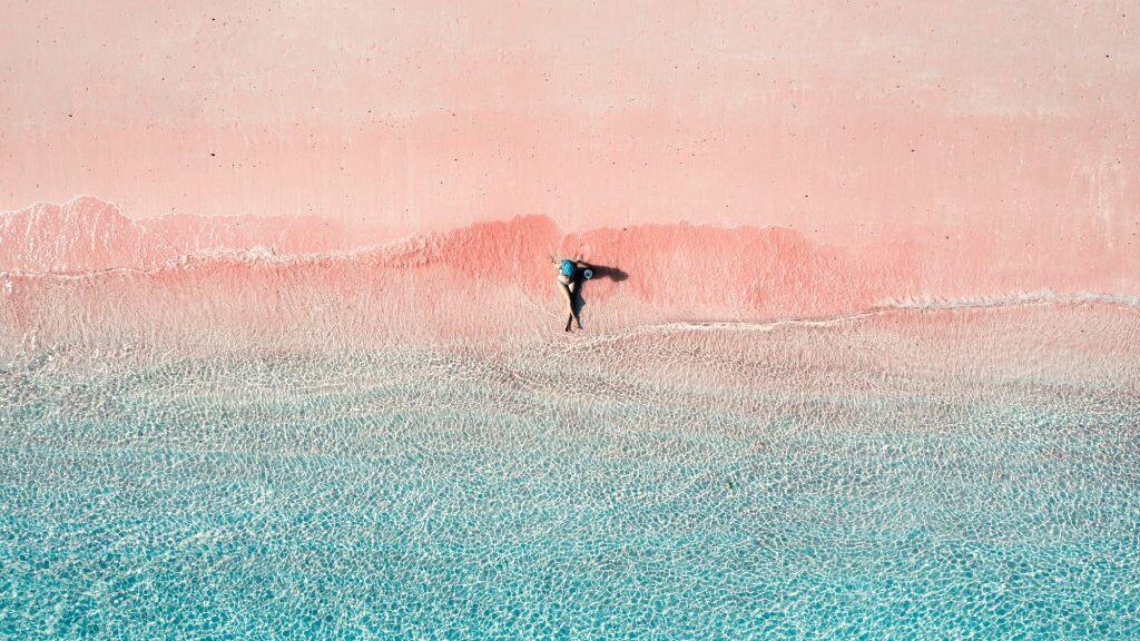 A person in a blue hat relaxes where gentle turquoise waves meet the soft pink sand, creating a dreamy, pastel-toned beachscape viewed from above.