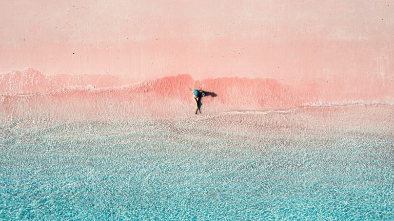 A person in a blue hat relaxes where gentle turquoise waves meet the soft pink sand, creating a dreamy, pastel-toned beachscape viewed from above.
