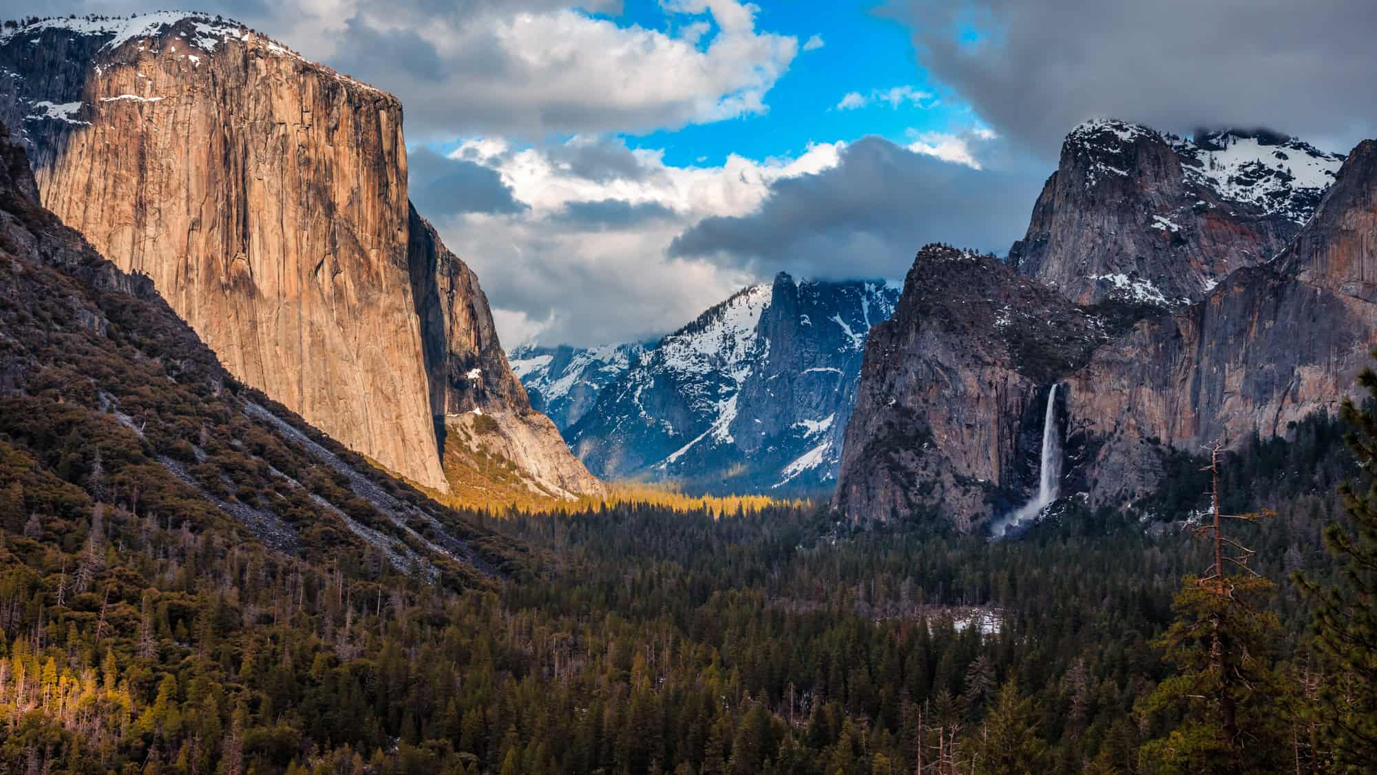 The famous view from Tunnel View showing El Capitan, Bridalveil Fall, and Half Dome, surrounded by pine forests and dramatic, snow-dusted peaks under a partly cloudy sky.