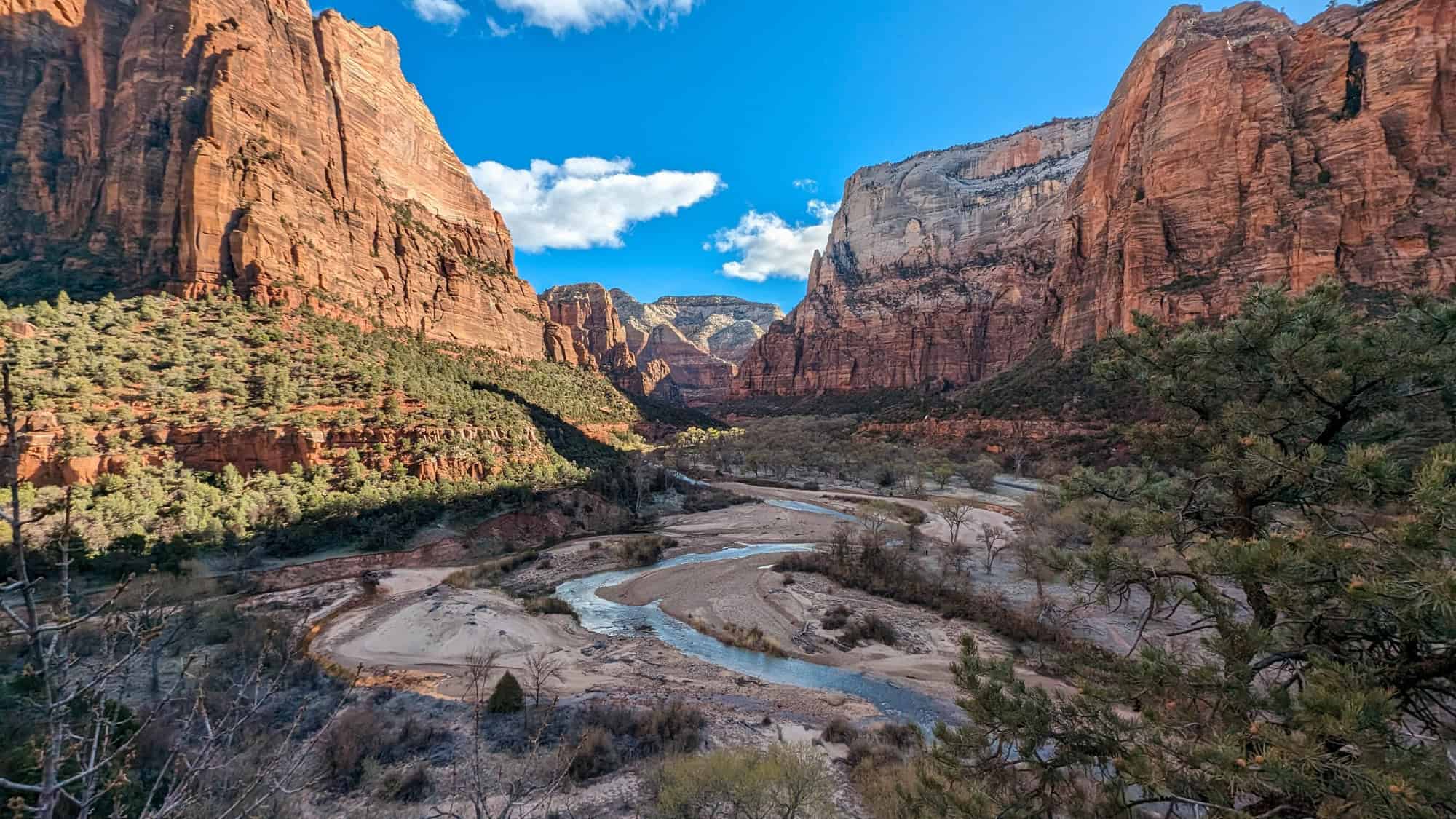 Majestic red rock cliffs rise high above a winding river and valley floor covered in patches of green trees under a bright blue sky with scattered clouds.