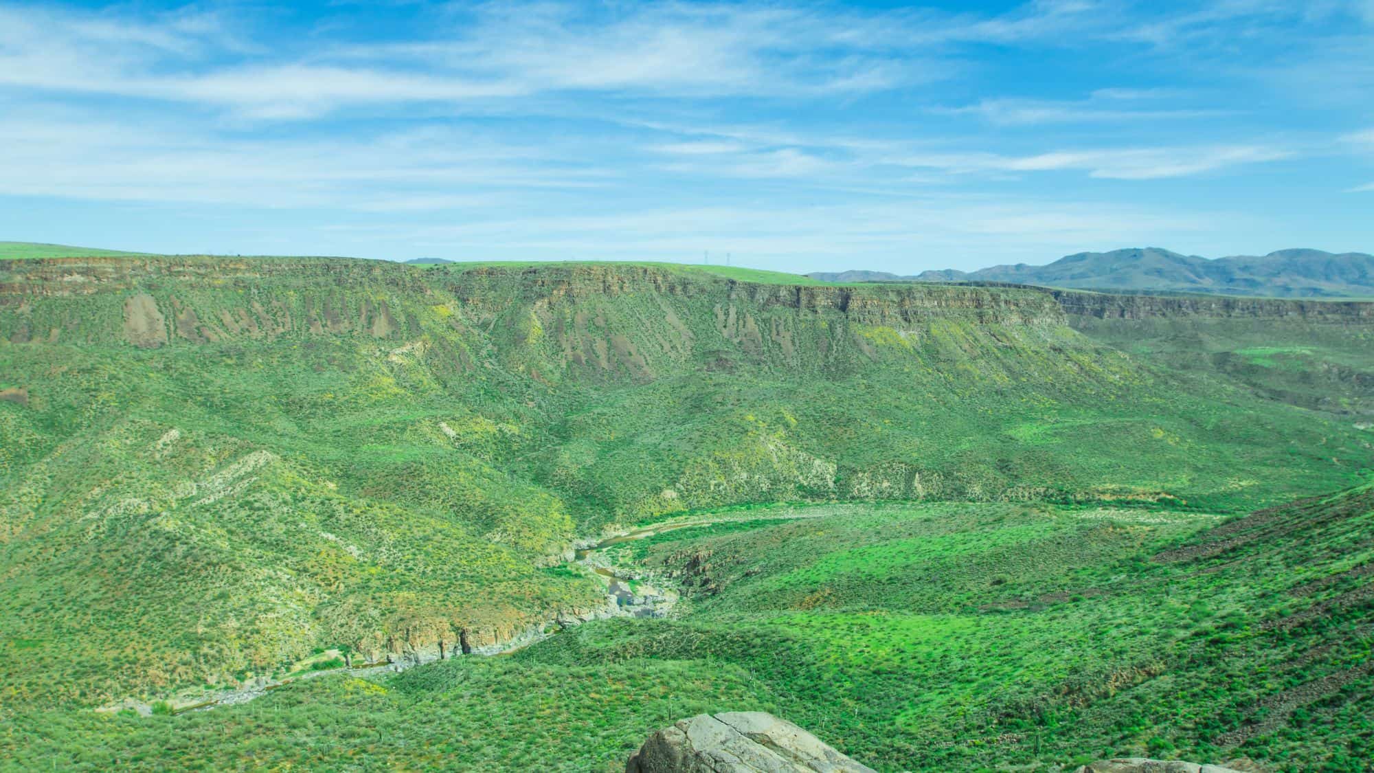 A lush, green canyon with rolling hills and a winding river under a bright blue sky.