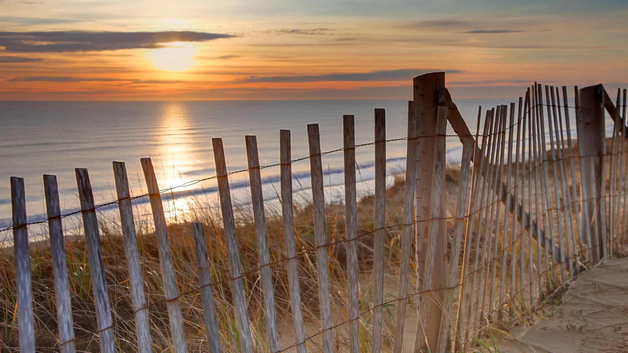 A serene sunset view from Cape Cod, Massachusetts, featuring golden sunlight reflecting off the ocean and a rustic wooden fence running along sandy dunes.