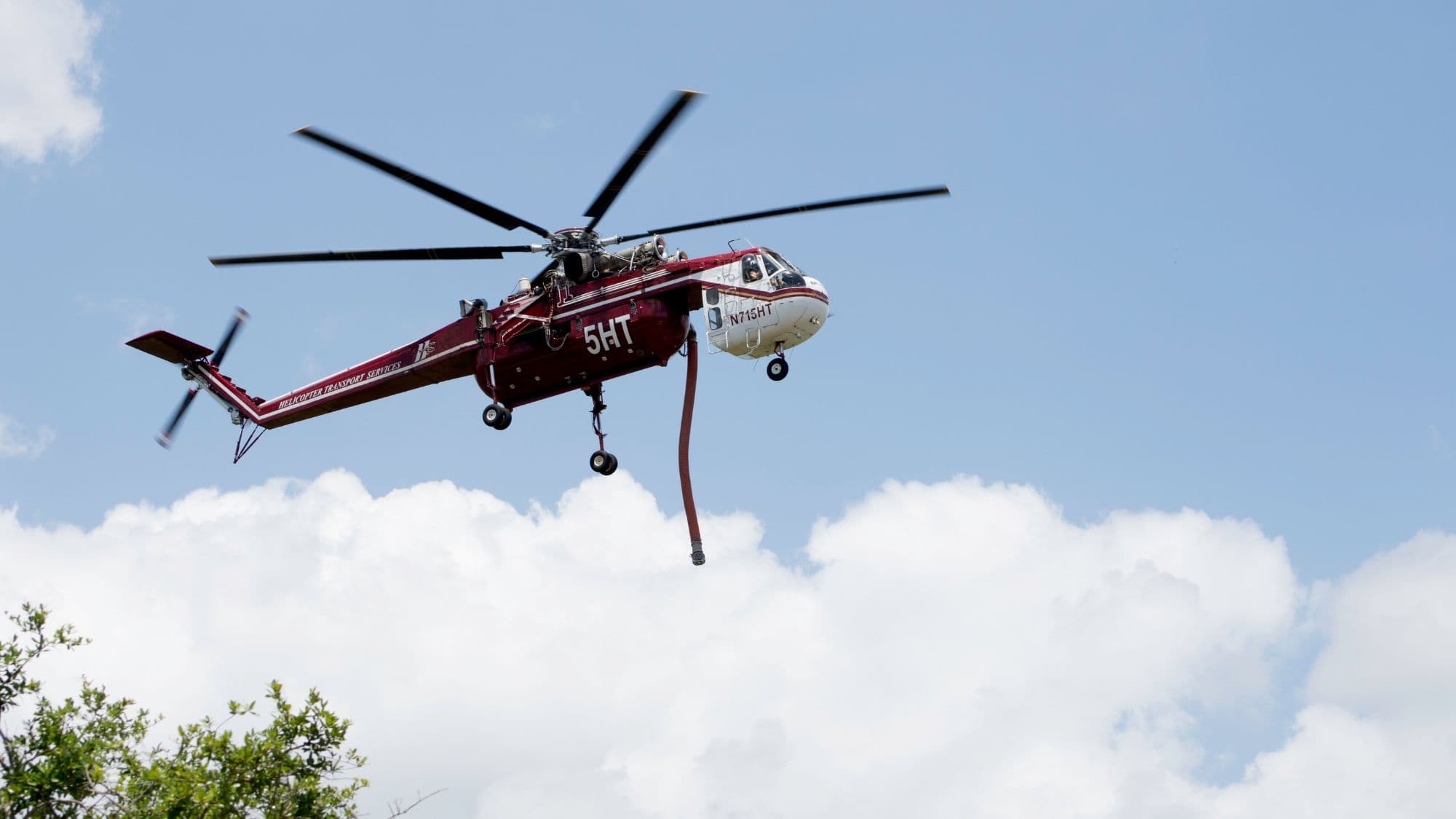 A red firefighting helicopter equipped with a water tank flies above a forested area, captured mid-air with a backdrop of fluffy white clouds.