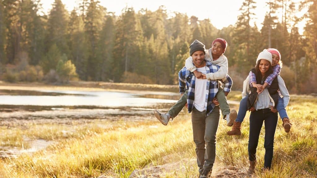 A family of four enjoys a hike through a serene forest by a lake, with the children happily riding piggyback, emphasizing outdoor fun and togetherness.