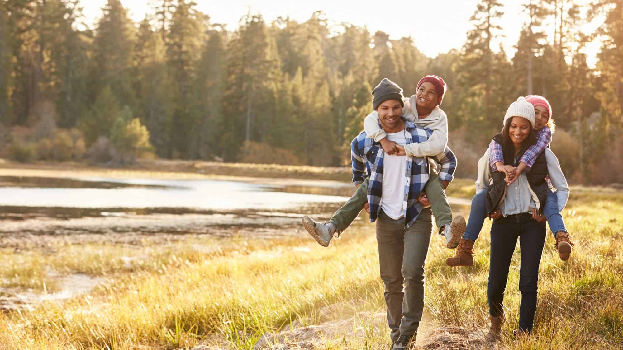 A family of four enjoys a hike through a serene forest by a lake, with the children happily riding piggyback, emphasizing outdoor fun and togetherness.

