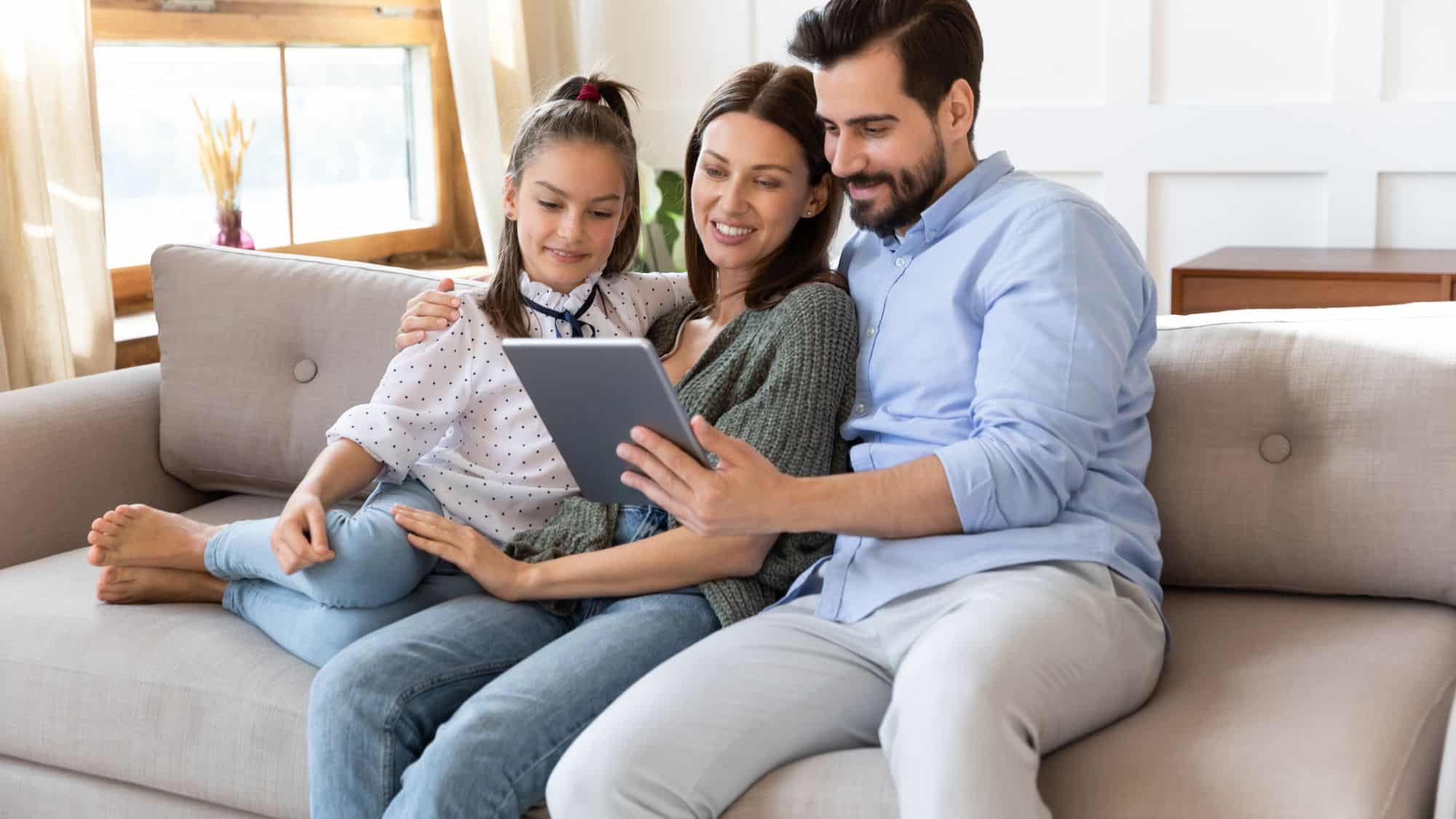 A cheerful family of three sitting on a cozy couch, smiling while looking at a tablet together. The scene captures a moment of shared bonding and digital interaction.