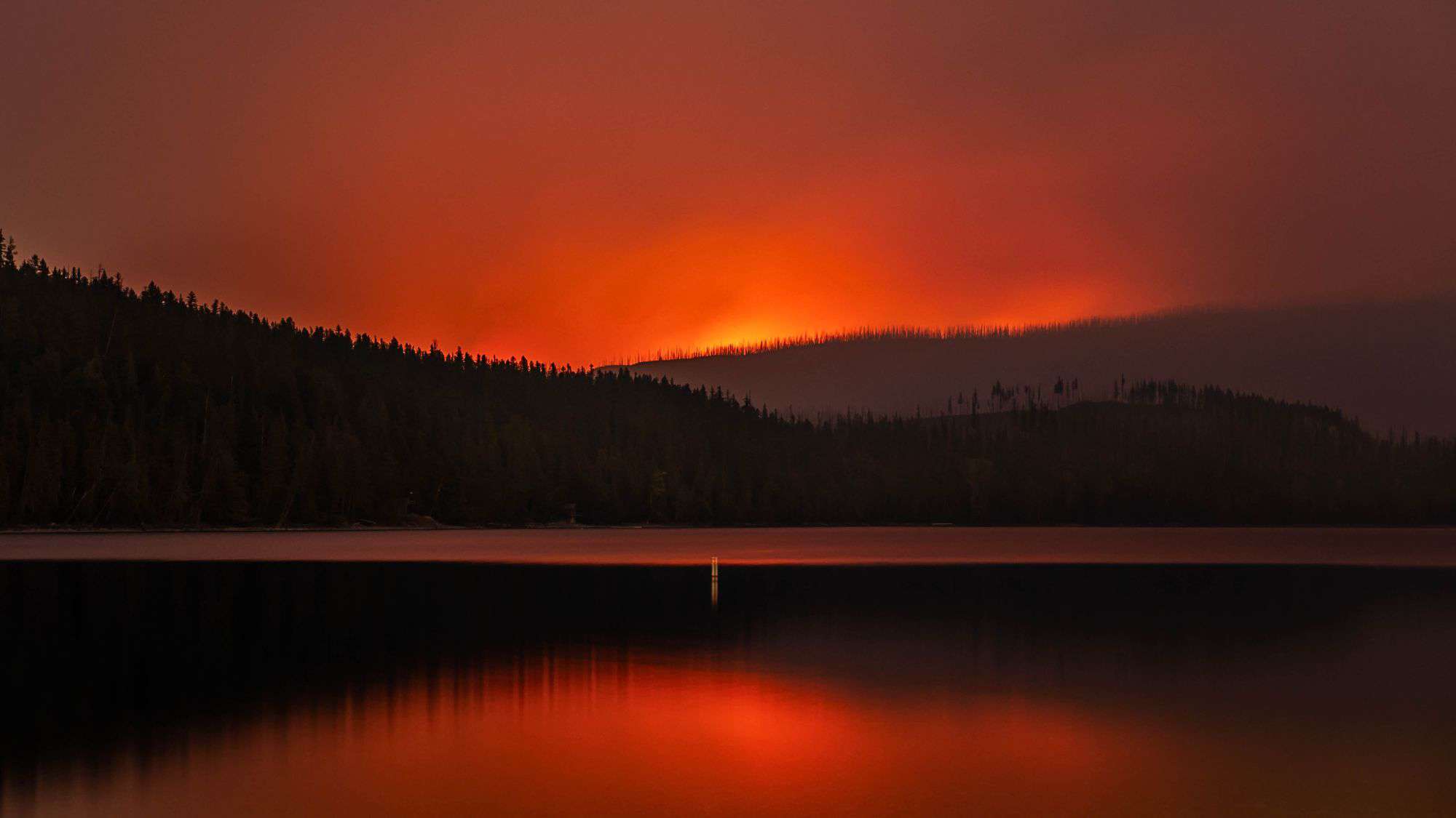 A dramatic image of an orange-red glow from a wildfire illuminating the sky over a silhouetted forest and a calm lake, with the fiery reflection shimmering on the water's surface.