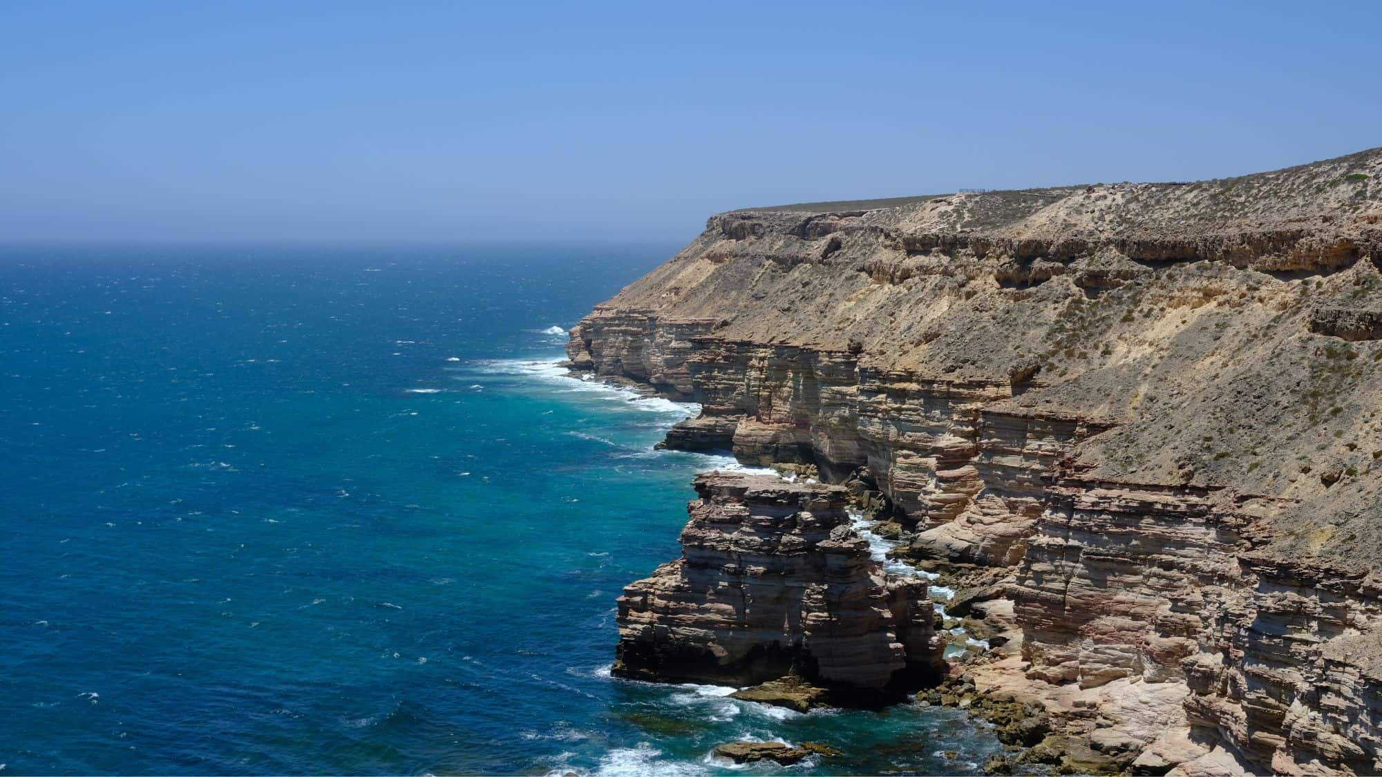 A vast coastal cliff with layers of red and white rock overlooking the deep blue sea. The rugged terrain contrasts sharply with the calm ocean below.