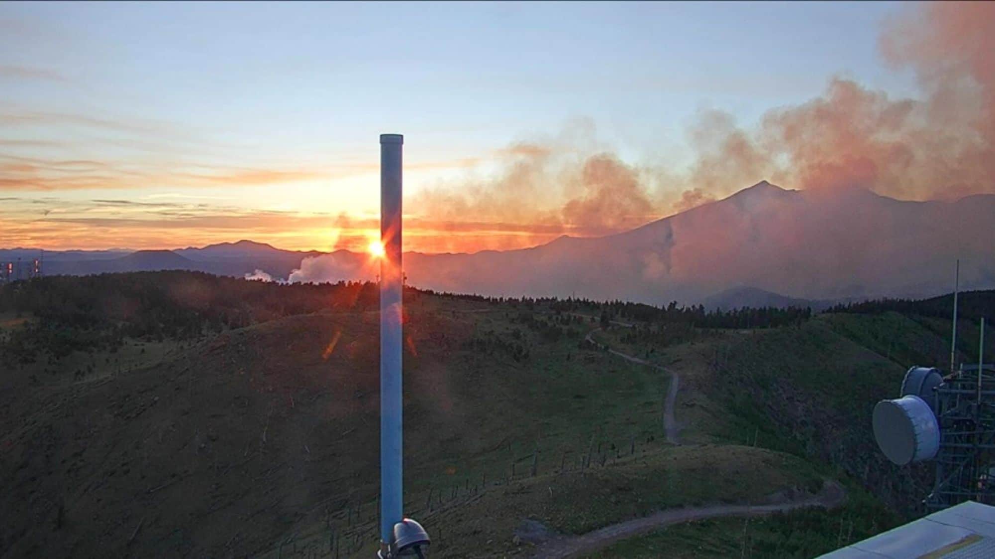 A sunset view of smoke rising from the Museum Fire, with the vibrant orange hues of the setting sun mixing with plumes of smoke against the distant mountains.