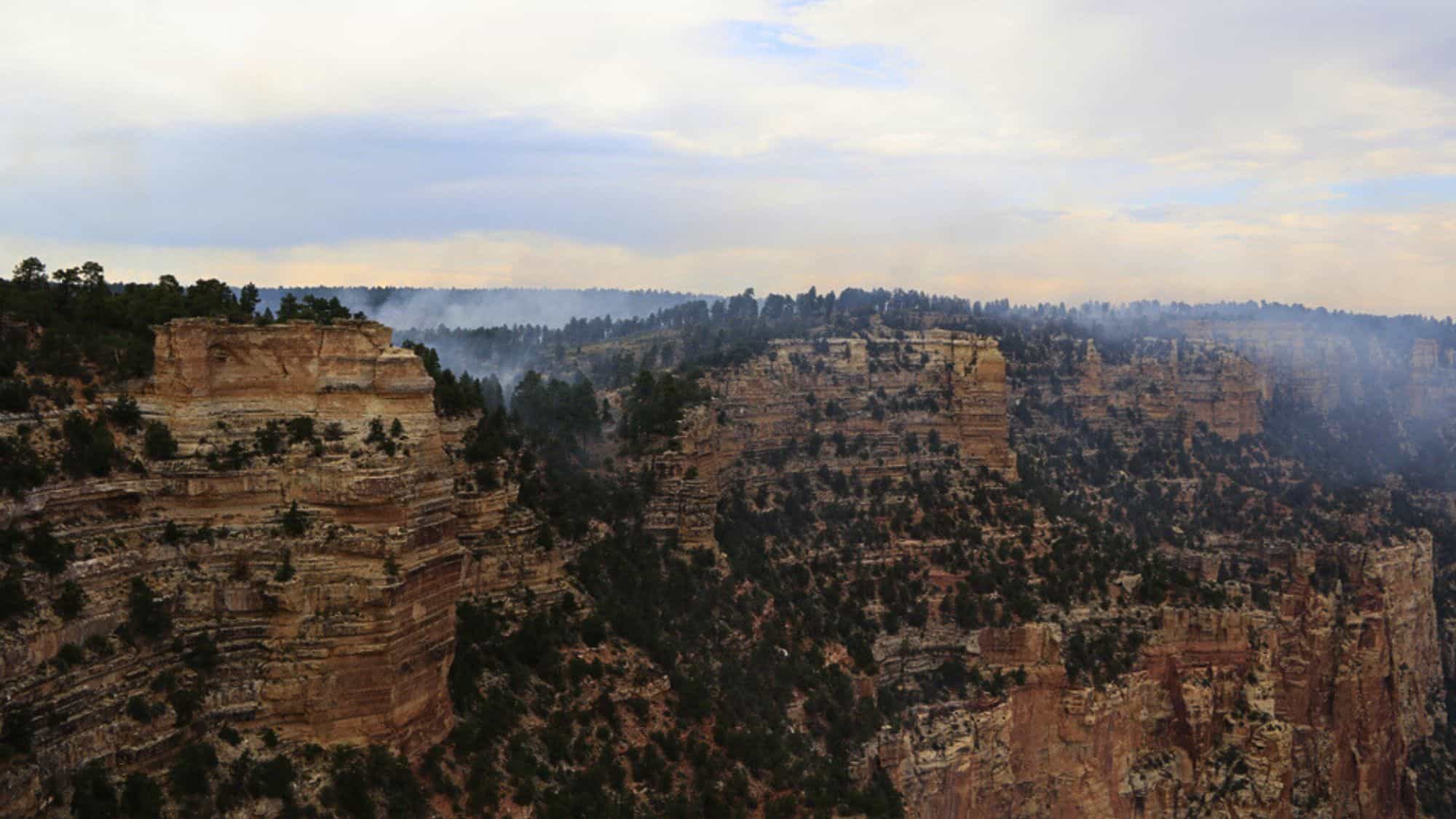 Smoke wafts over the cliffs and trees of the Grand Canyon’s North Rim, with the layered rock formations partially shrouded in haze.