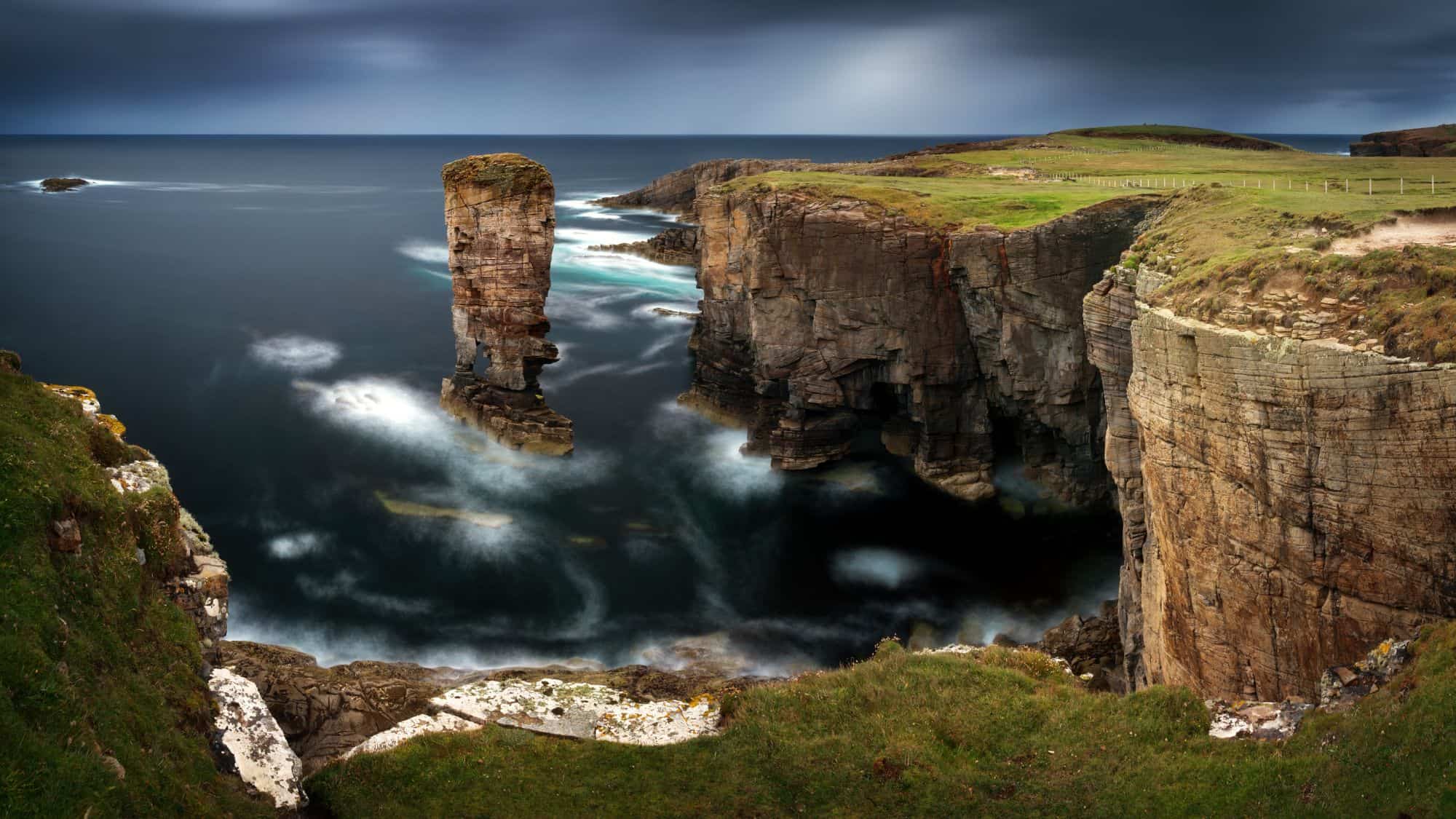 A dramatic coastal landscape featuring a towering sea stack surrounded by rugged cliffs and dark, turbulent waters. The overcast sky adds a sense of mystery and awe.