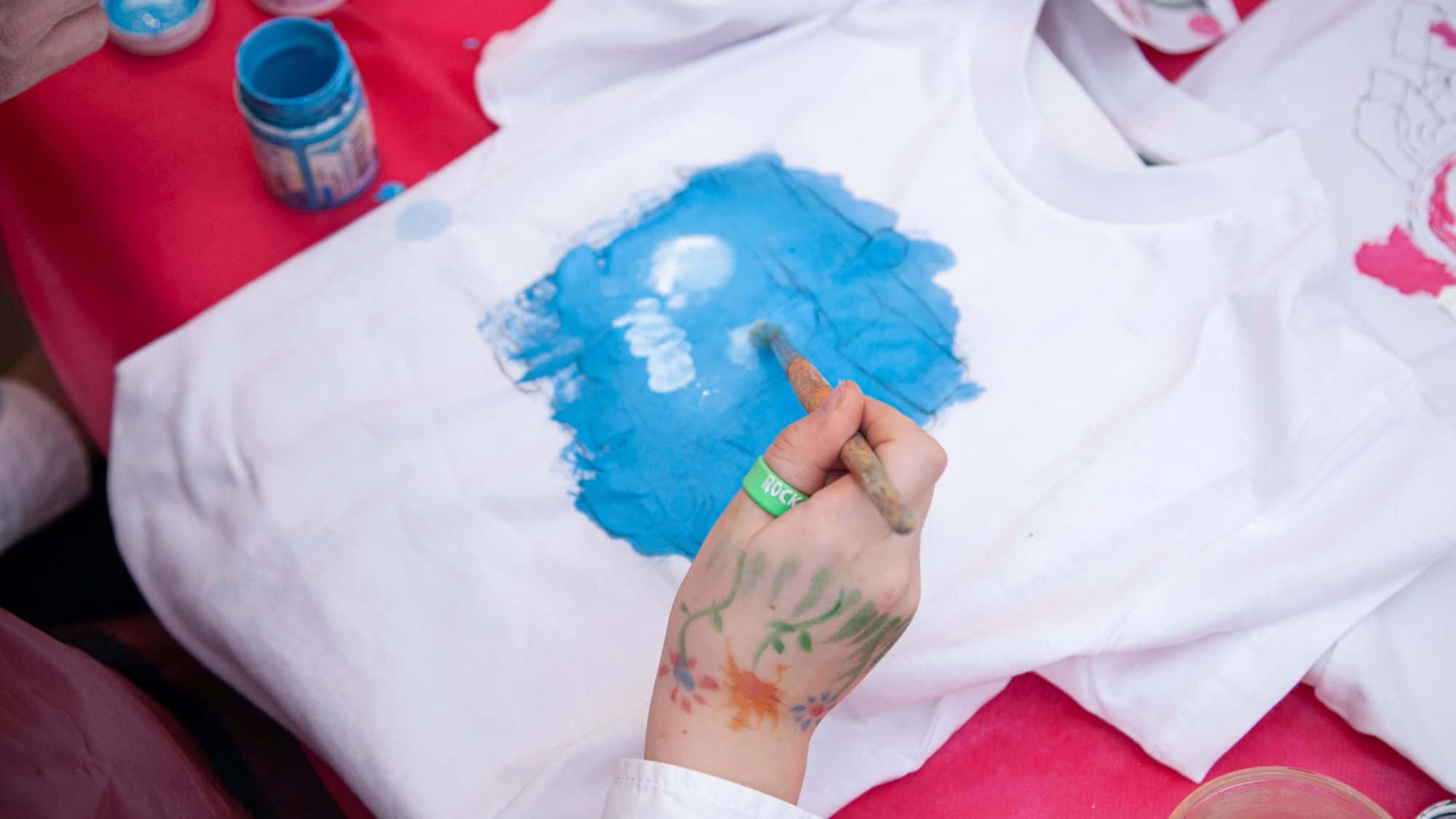 A close-up of a child's hand painting a design on a white t-shirt with bright blue paint. The child's hand features colorful hand-drawn tattoos, and paint jars are visible on a red surface.