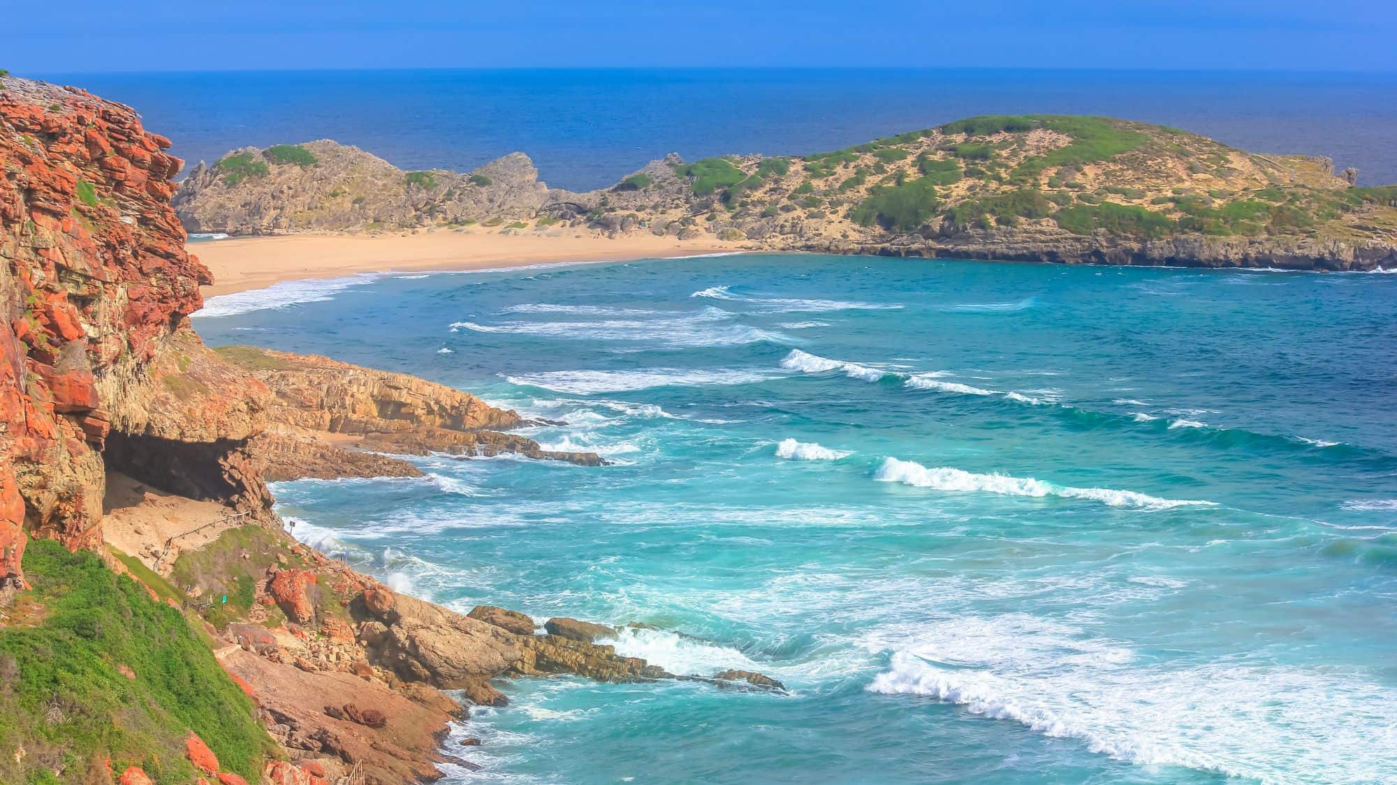 A vivid view of a turquoise ocean meeting golden sands, bordered by rocky cliffs covered in greenery. The rolling waves add dynamic energy to the picturesque scene.