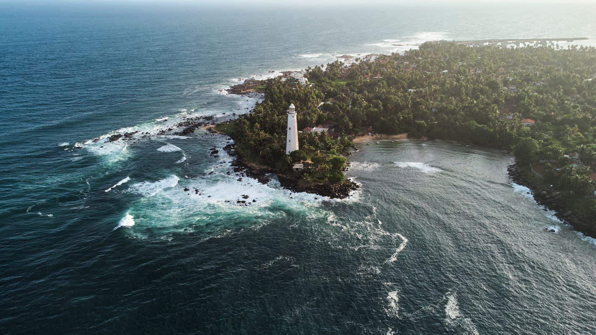 An aerial view of a lighthouse perched on a lush, green peninsula surrounded by vibrant blue ocean waves crashing against rocky shores.