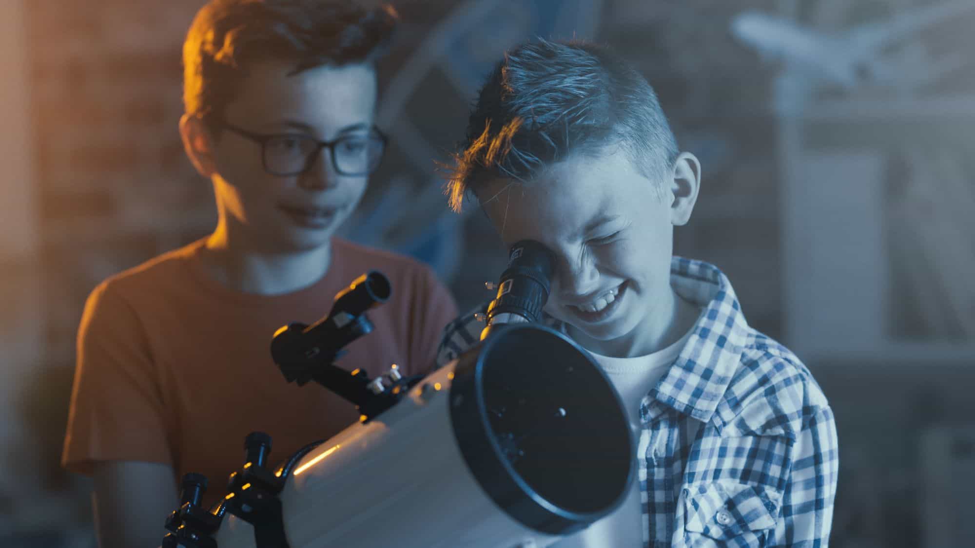 Two boys exploring the night sky with a telescope indoors. One boy peers through the telescope while the other looks on, reflecting curiosity and wonder in a dimly lit setting.