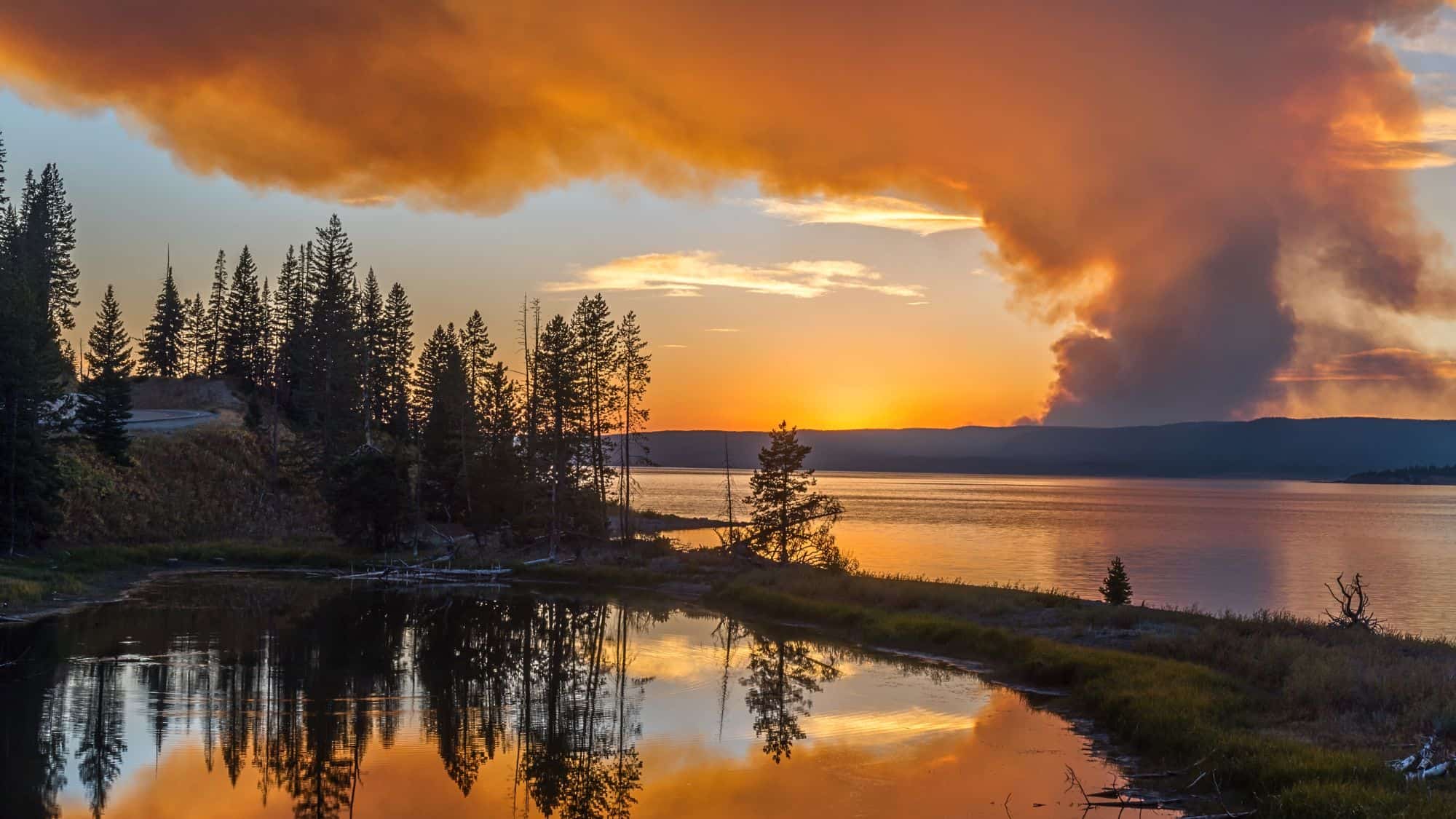 A picturesque scene of a fiery orange and red sunset over a serene lake, with billowing smoke from a wildfire rising in the distance, framed by pine trees reflecting in a small pond.