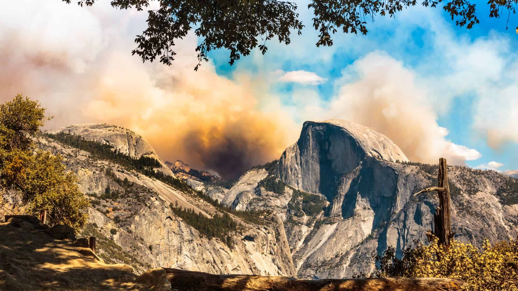 Smoke drifts over Yosemite’s iconic Half Dome and surrounding cliffs, with a mix of blue skies and brownish haze from an active wildfire in the area.