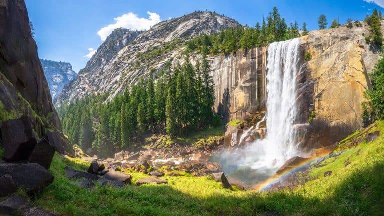 A panoramic view of a stunning waterfall cascading down a granite cliff, surrounded by lush greenery and tall pine trees, with a rainbow forming at the base.