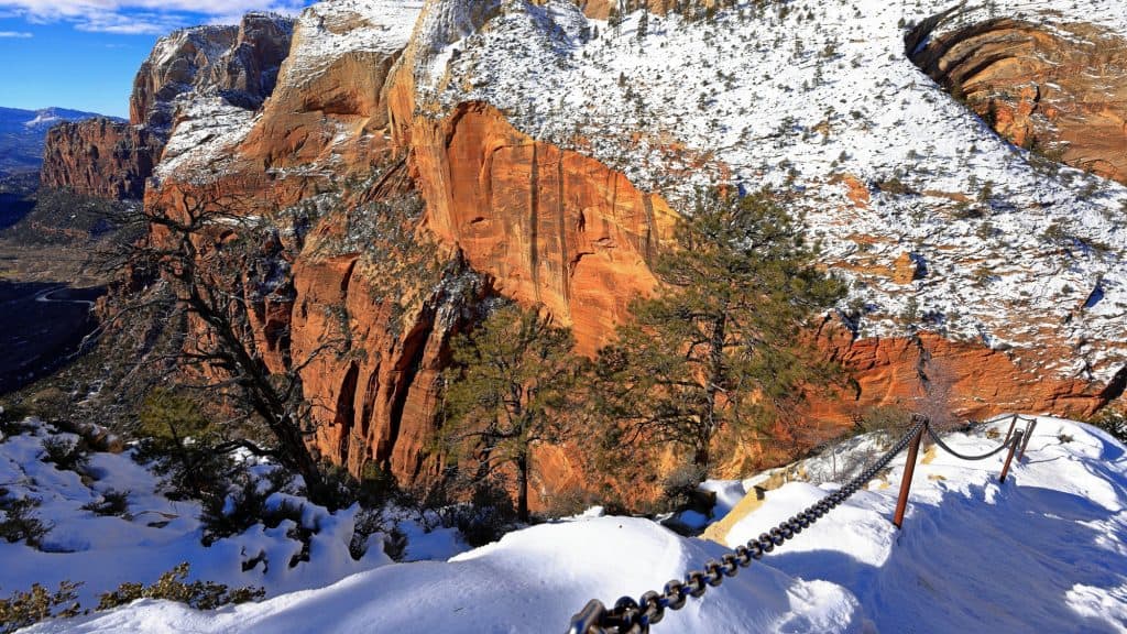 A snowy winter landscape of Zion National Park featuring steep red sandstone cliffs adorned with patches of snow and sparse evergreen trees. A metal chain railing stretches diagonally across the foreground, marking a snowy pathway. The background reveals a winding river or road in the canyon below, with a clear blue sky and wispy clouds above.