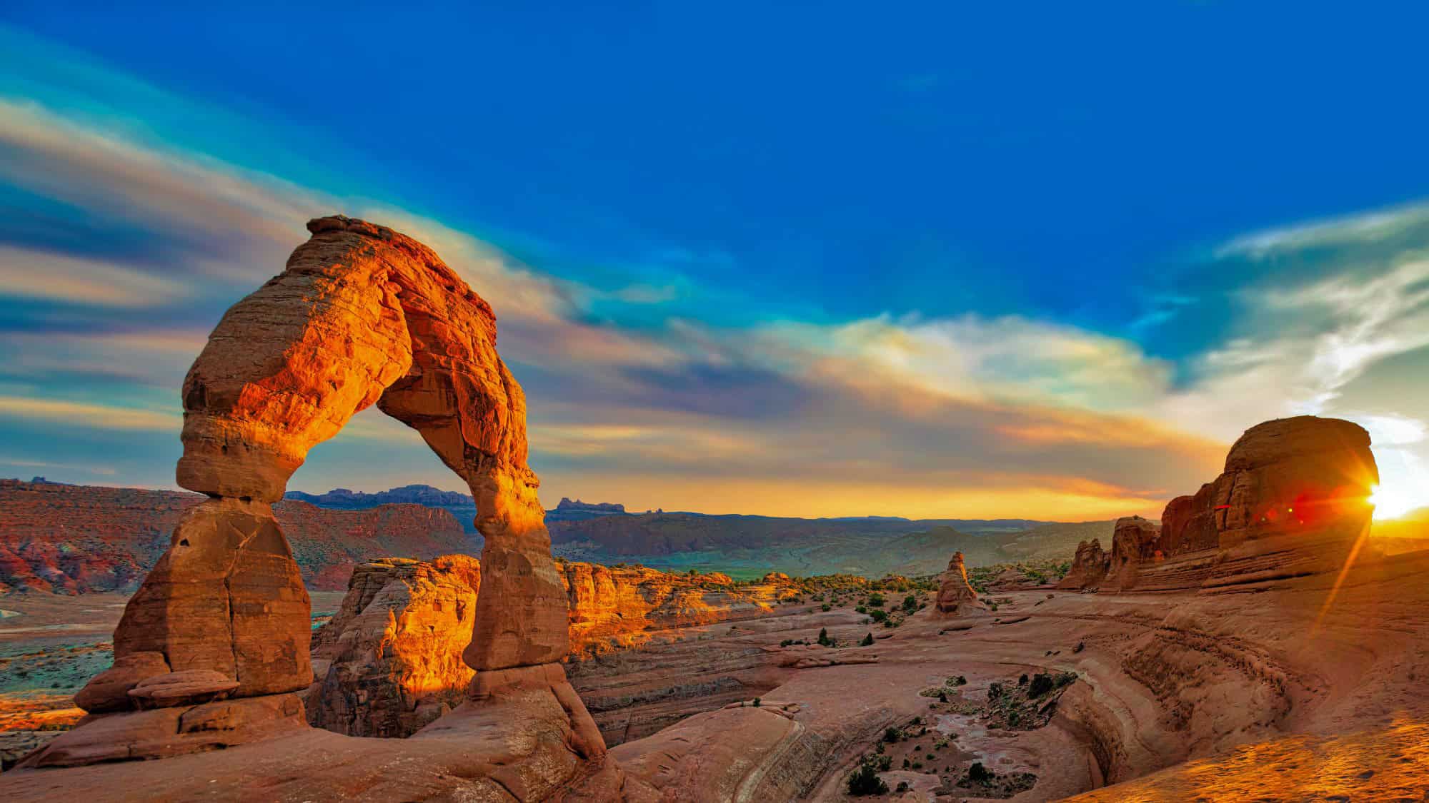 The iconic Delicate Arch glowing in golden tones as the sun sets, painting the sky with a blend of warm colors over the desert landscape.