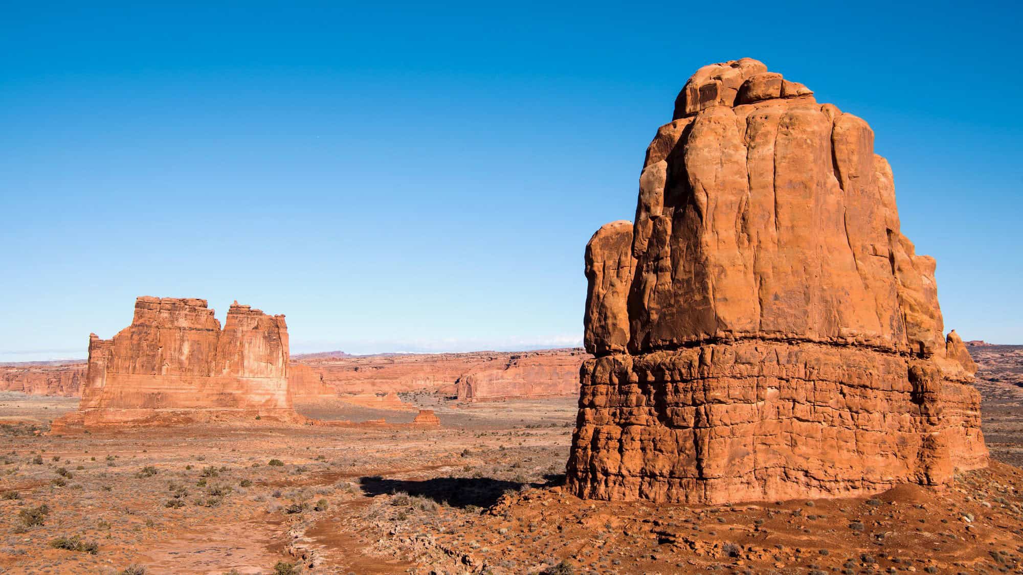 Massive red rock monoliths and mesas rising against a vivid blue sky, with expansive desert plains stretching in the foreground.