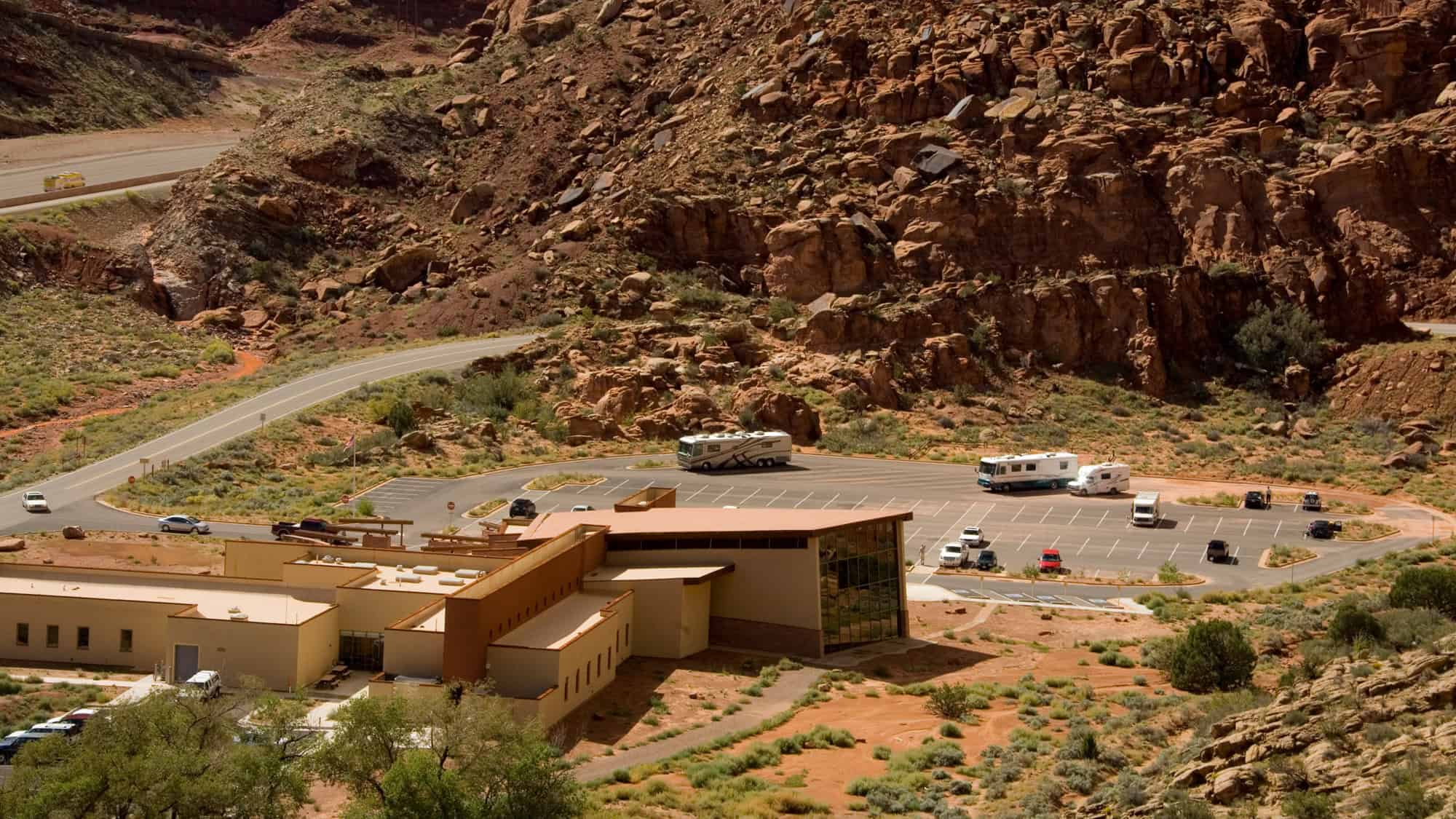 The modern visitor center nestled against a rocky hillside, with ample parking and desert flora surrounding the structure.