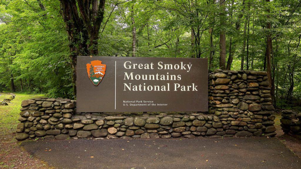 A stone and wood sign marks the entrance to Great Smoky Mountains National Park, set in a lush green forest with moss-covered rocks and towering trees.
