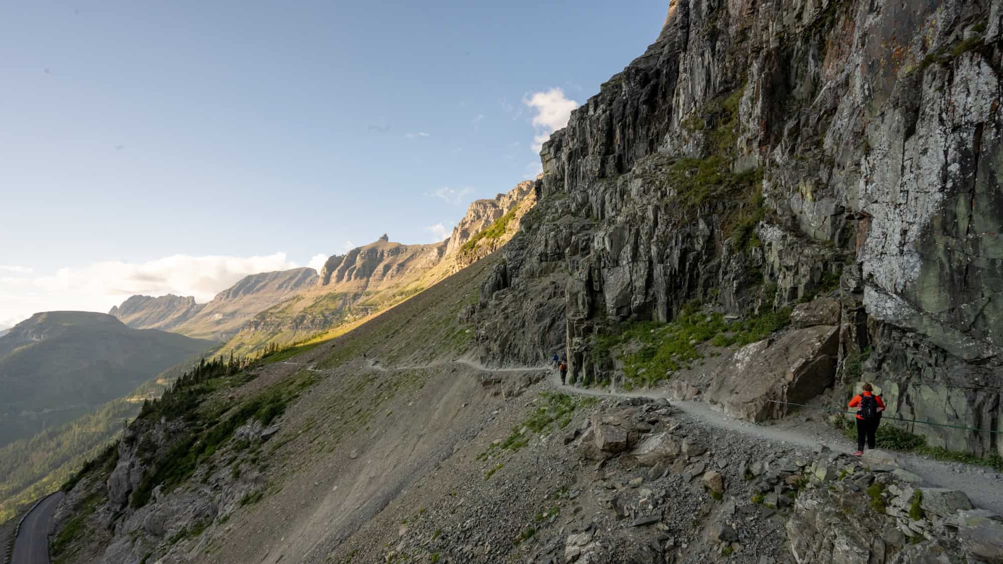 A narrow dirt trail winds along the side of a steep, rocky mountain face under a bright sky. A hiker in an orange jacket walks along the trail near a rope barrier, with other hikers visible in the distance. The rugged landscape features towering rock formations, sparse vegetation, and distant mountain ranges fading into atmospheric haze.