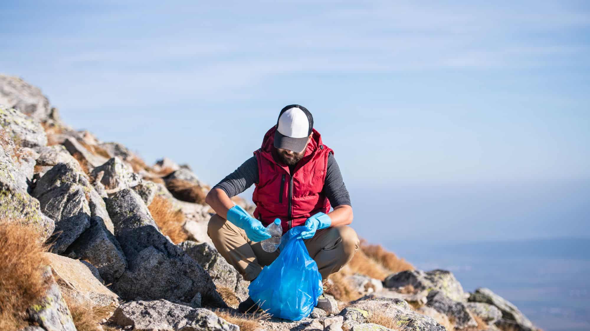 A man wearing a red vest and gloves is crouched on a rocky mountain path, cleaning up litter with a blue trash bag, contributing to environmental preservation.