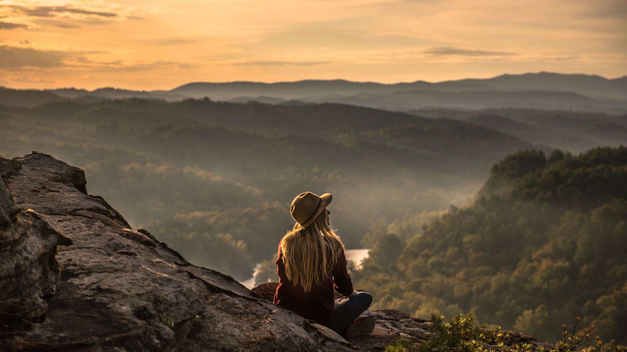 A woman in a wide-brimmed hat sits on a rocky ledge, gazing at a serene valley below bathed in the golden glow of sunset.