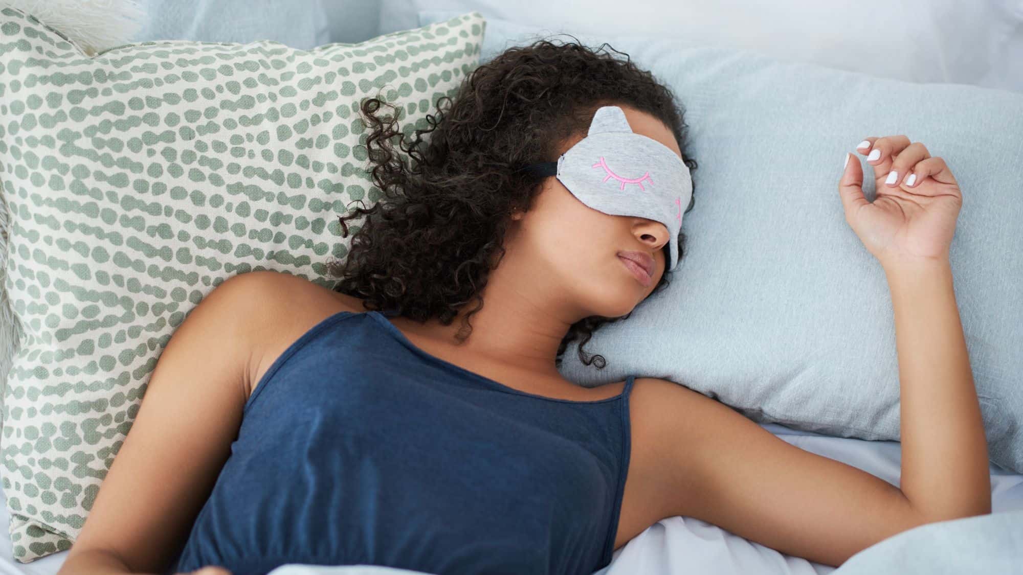 A woman sleeps peacefully in bed wearing a gray eye mask with a pink cat face design, surrounded by soft, patterned pillows.