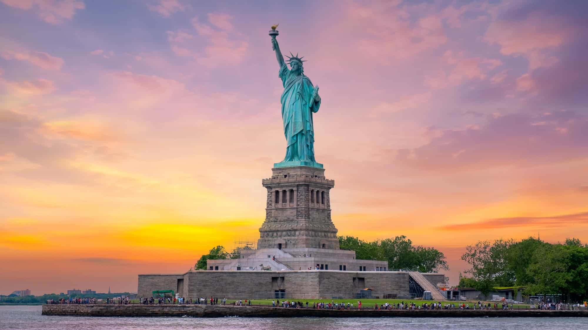 The Statue of Liberty is silhouetted against a vibrant sunset sky, with hues of orange, pink, and purple, creating a serene and majestic scene.