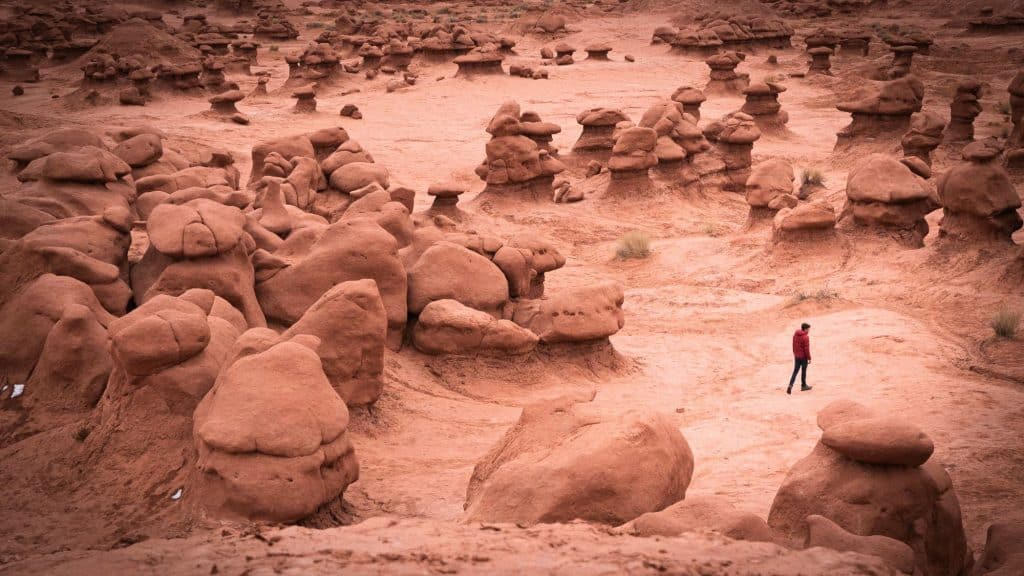 A surreal desert landscape filled with unique, mushroom-shaped rock formations, known as hoodoos, at Goblin Valley State Park in Utah. A lone hiker in a red jacket walks among the oddly shaped rocks, adding a sense of scale to the alien-like terrain.