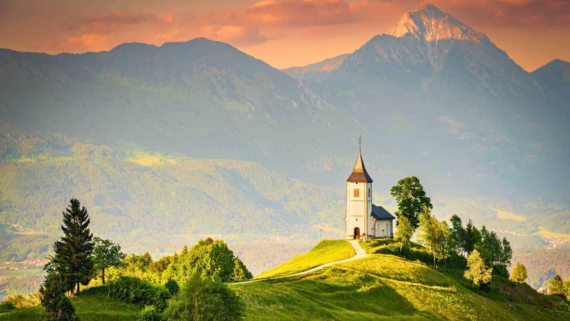 A charming white church with a pointed steeple sits atop a rolling green hill, bathed in the golden glow of sunset. Behind it, a backdrop of majestic mountains stretches across the horizon, partially covered in soft mist.