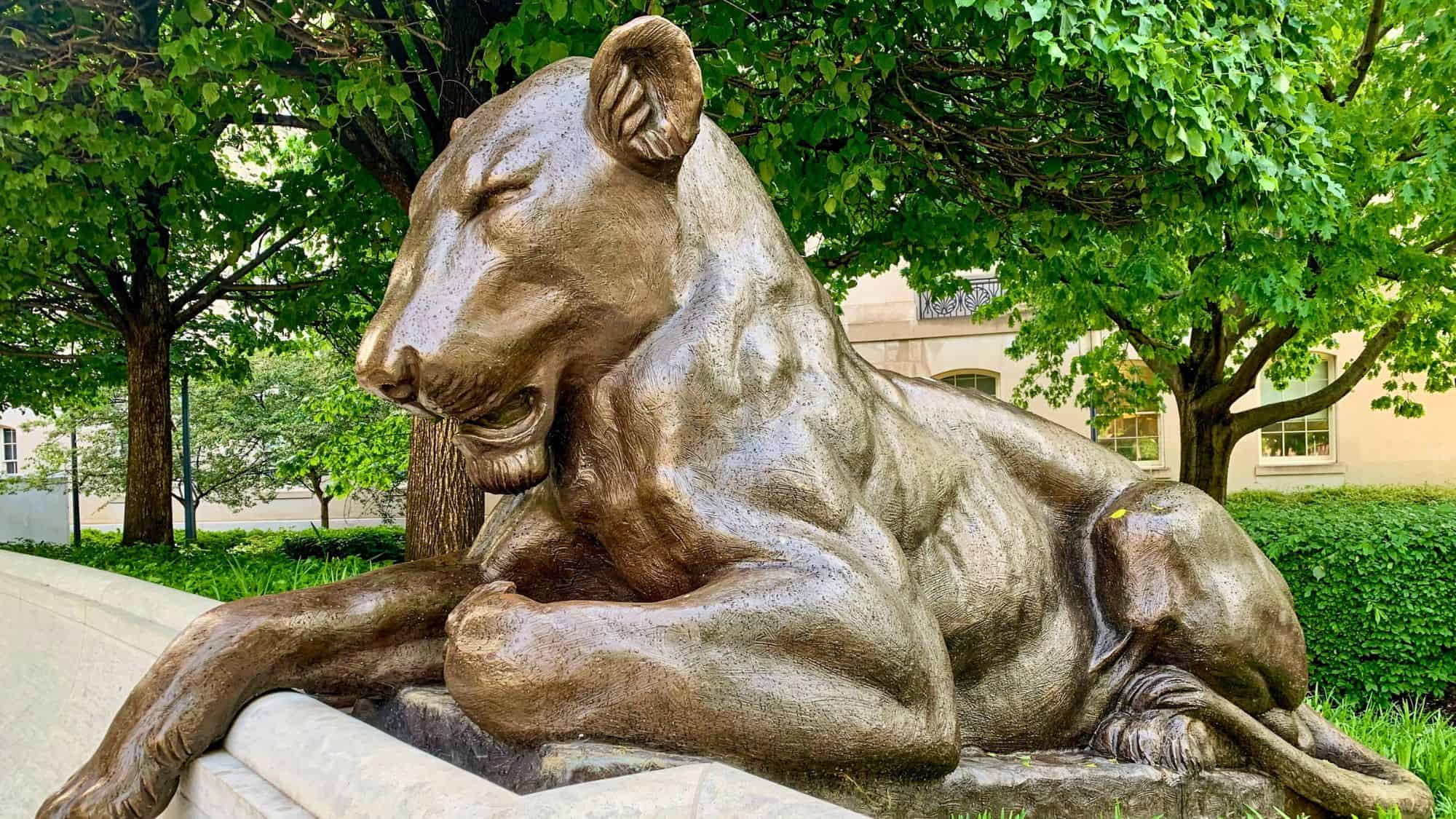 A bronze lion statue rests calmly atop a stone base, its muscular form softened by time and framed by leafy trees and a stately building in the background. The polished surface catches the light, giving the scene a quiet, dignified presence within a landscaped setting.