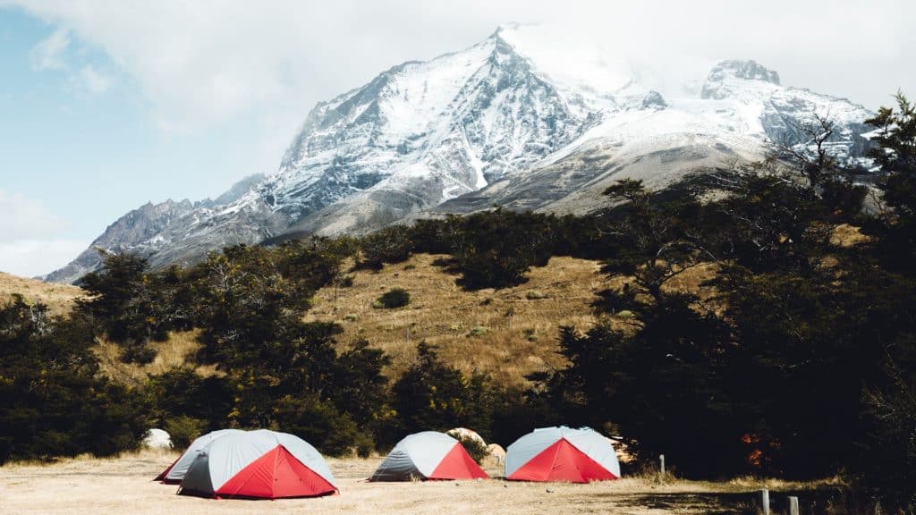 A campsite with several dome-shaped tents set up at the base of a towering, snow-capped mountain in Torres del Paine National Park, Chile. The rugged landscape, with dry grass and scattered trees, contrasts with the dramatic peaks in the background.