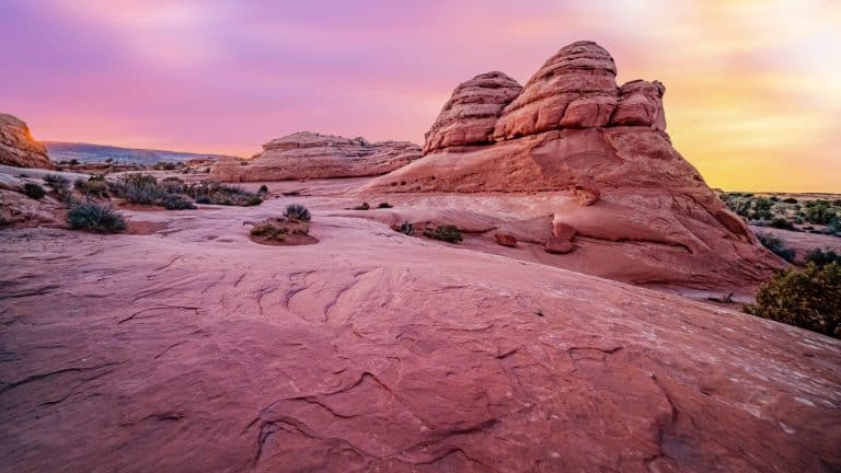 A peaceful desert landscape at sunset, with smooth, rounded sandstone formations glowing in shades of pink and orange. The sky transitions from soft purple to golden hues, creating a dreamy atmosphere. Sparse vegetation dots the arid terrain, enhancing the vast, open feel.
