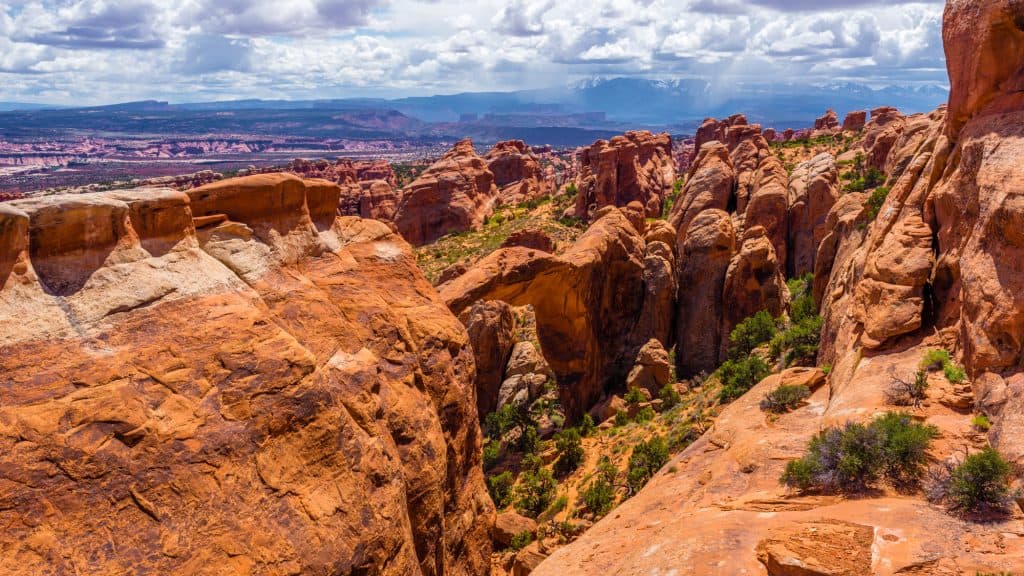 A stunning view of rugged sandstone formations with a natural arch spanning between red rock cliffs. The background reveals vast desert landscapes and distant mountains under a partly cloudy sky.