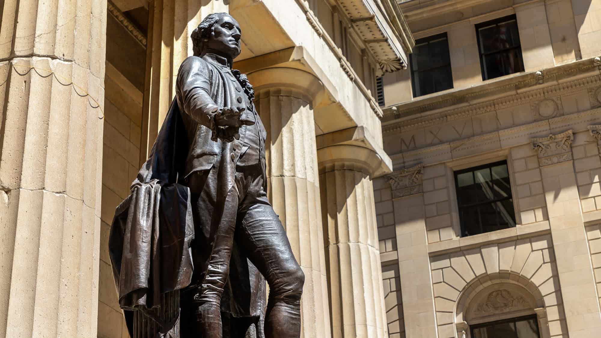A bronze statue of George Washington stands in front of a grand classical building with fluted columns, symbolizing the site of his first presidential oath.
