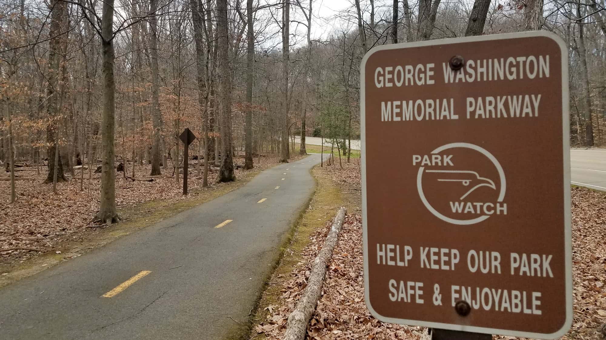 A brown sign with white text reading "George Washington Memorial Parkway" stands beside a paved trail in a forested area during autumn, surrounded by bare trees and fallen leaves.