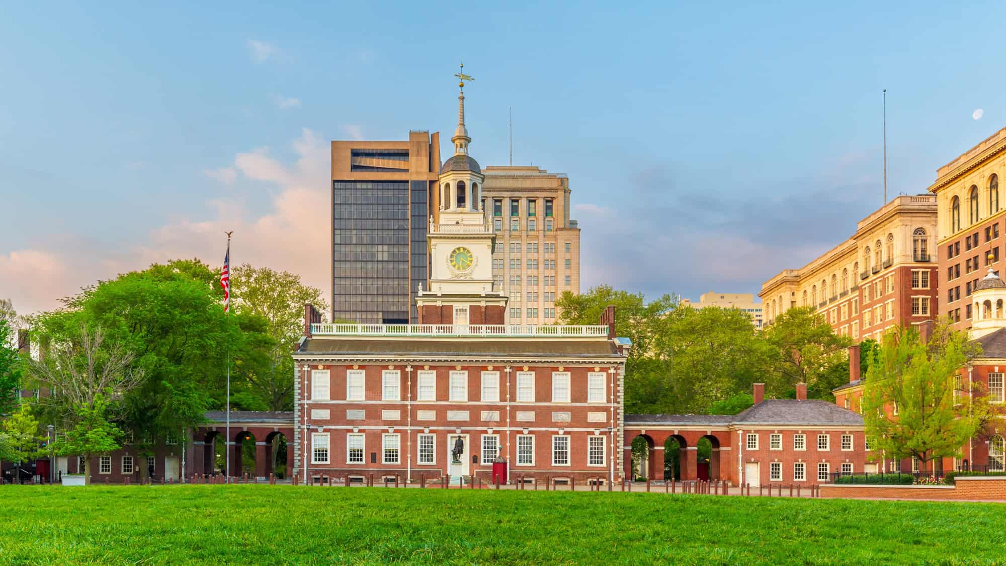 Independence Hall, a red brick colonial building with a white clock tower, is framed by lush green lawns and trees, set against a backdrop of modern skyscrapers.