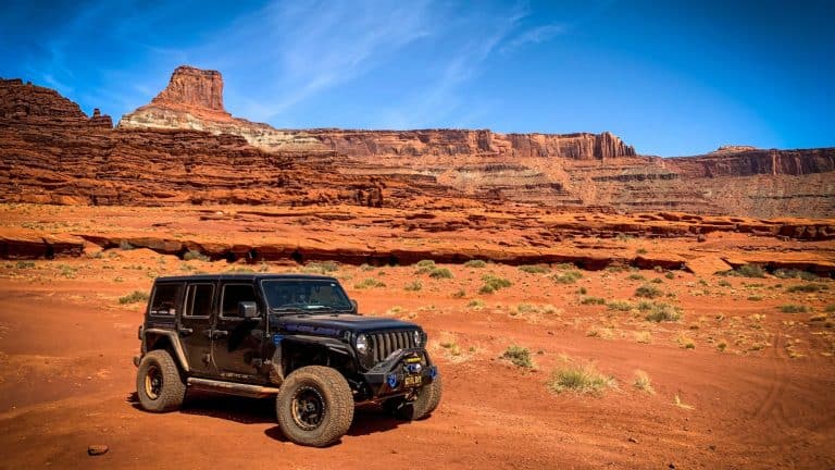 A black Jeep with off-road modifications is parked on the red sandy terrain of Moab, Utah, with towering red rock formations and a clear blue sky in the background.