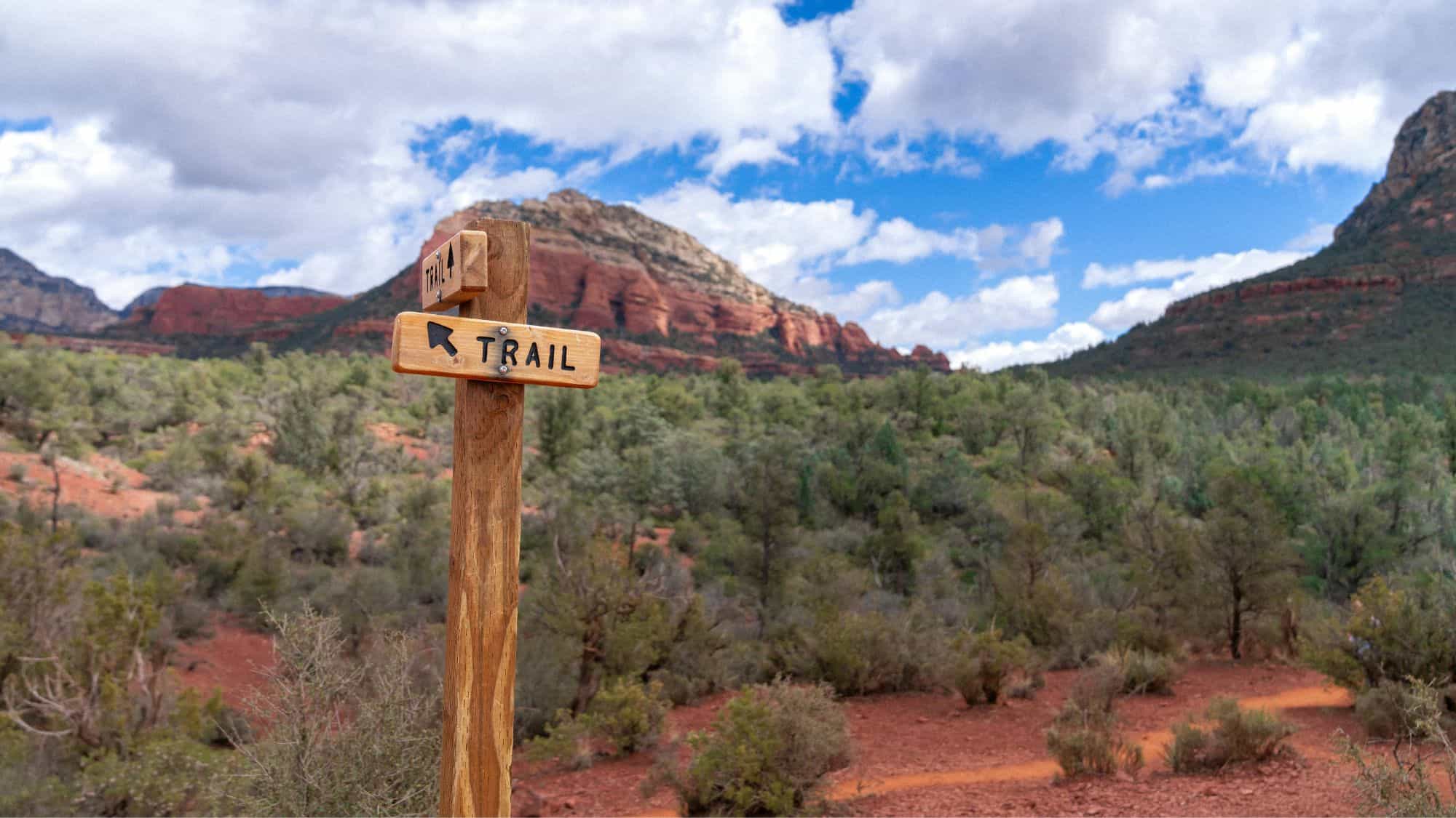 A rustic wooden trail marker with the word "TRAIL" and an arrow pointing left, surrounded by desert vegetation and red rock formations under a bright blue sky with scattered clouds.